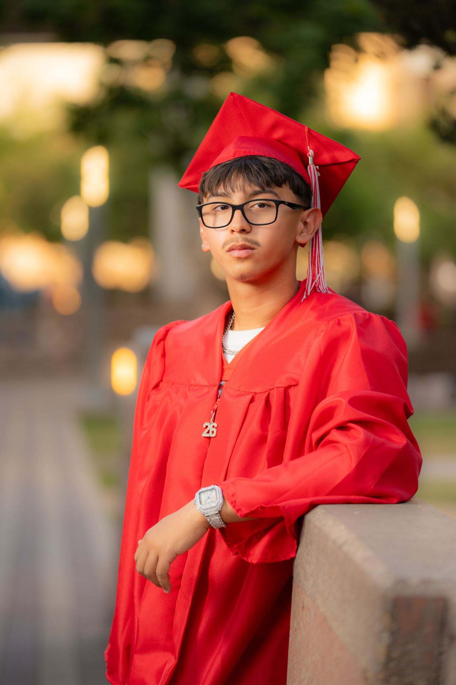Young man in red graduation cap and gown with glasses poses outdoors for senior portrait photo.