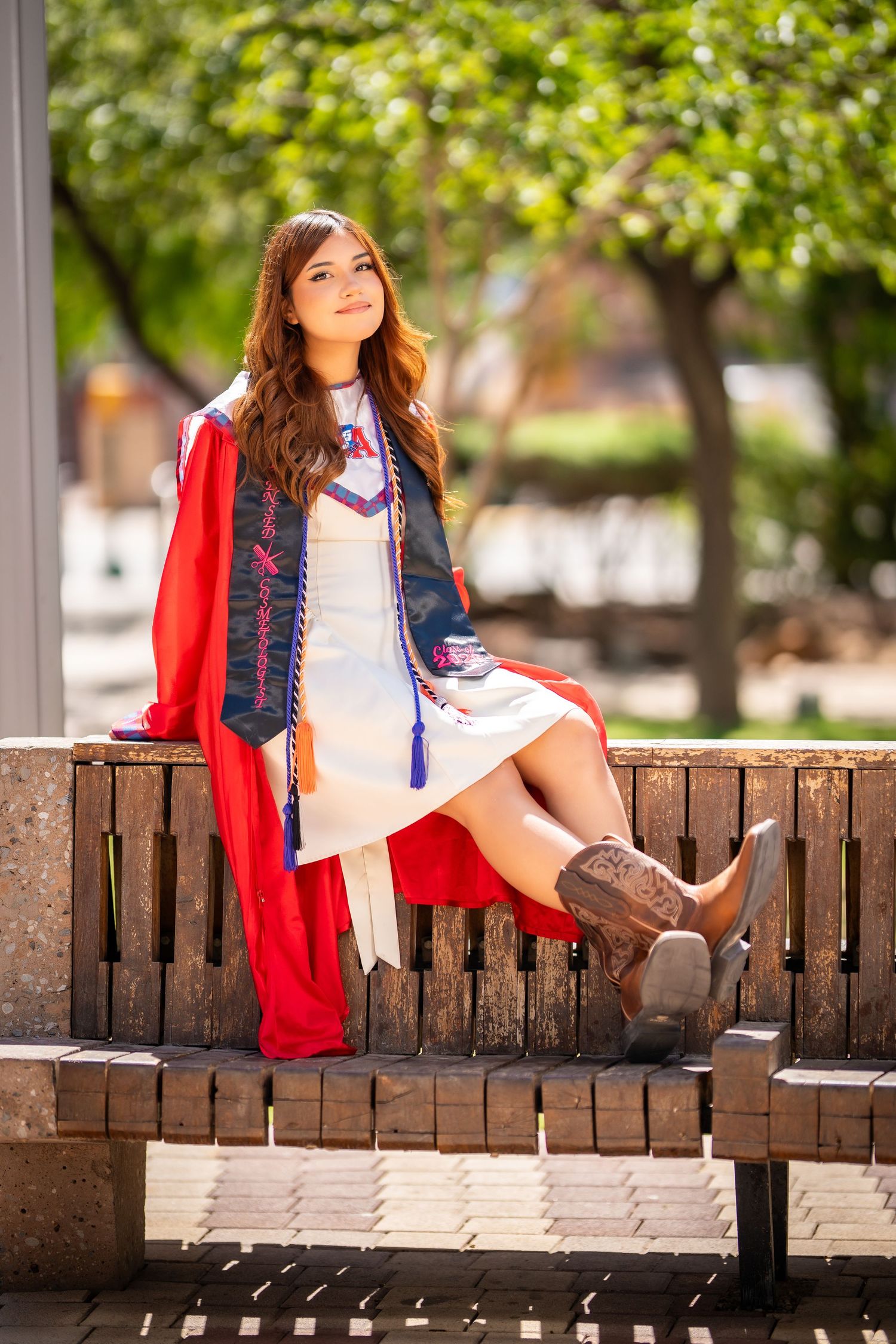 Young woman in red graduation gown and cowboy boots sitting on a bench outdoors, celebrating her graduation day.