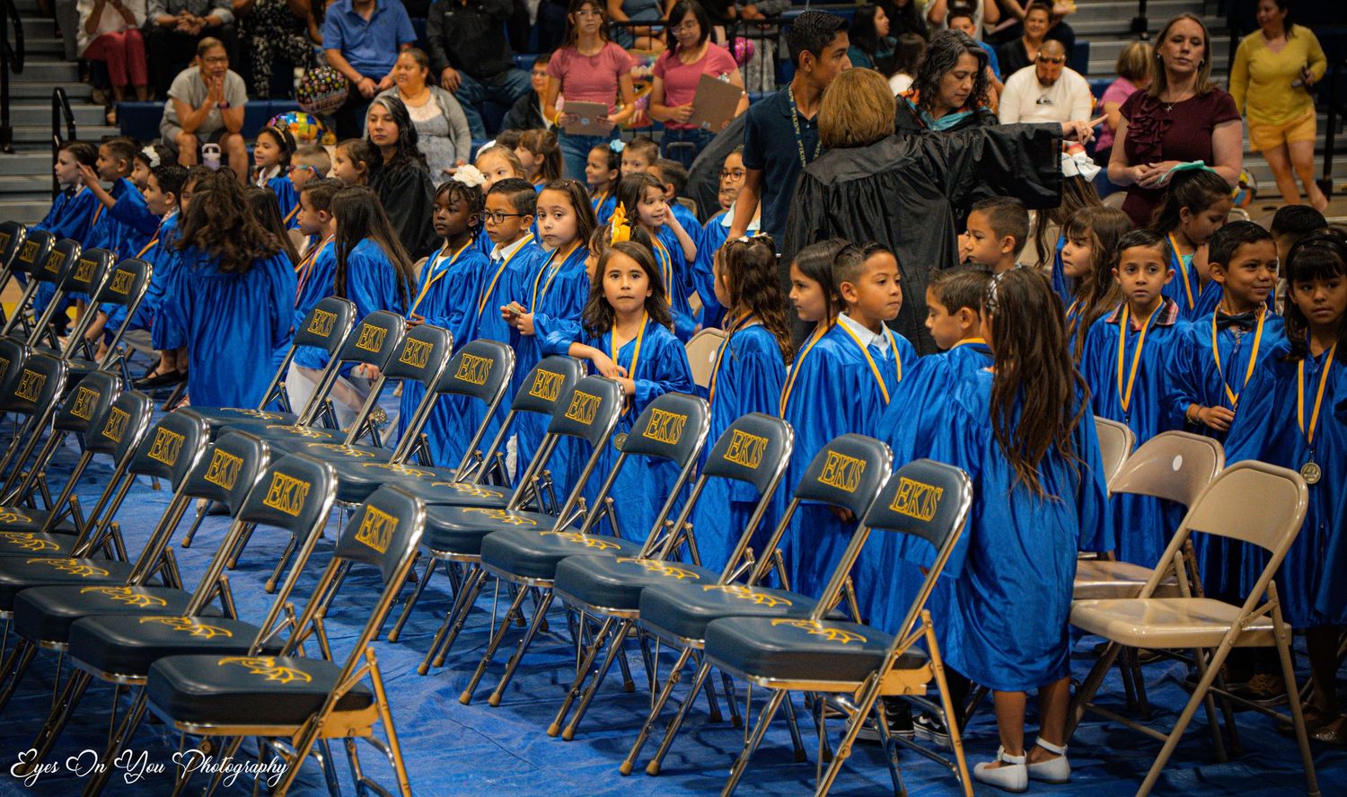 Audience seated in blue chairs during school ceremony in gymnasium.