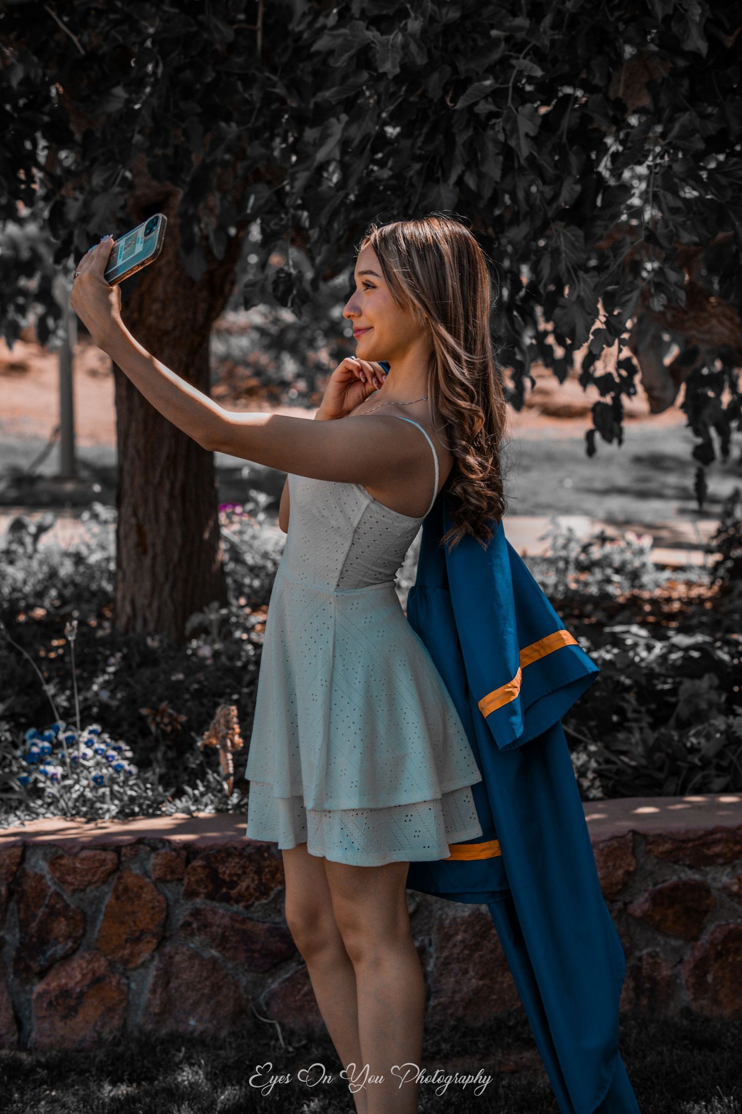 Graduate in white dress taking selfie while holding blue graduation gown.