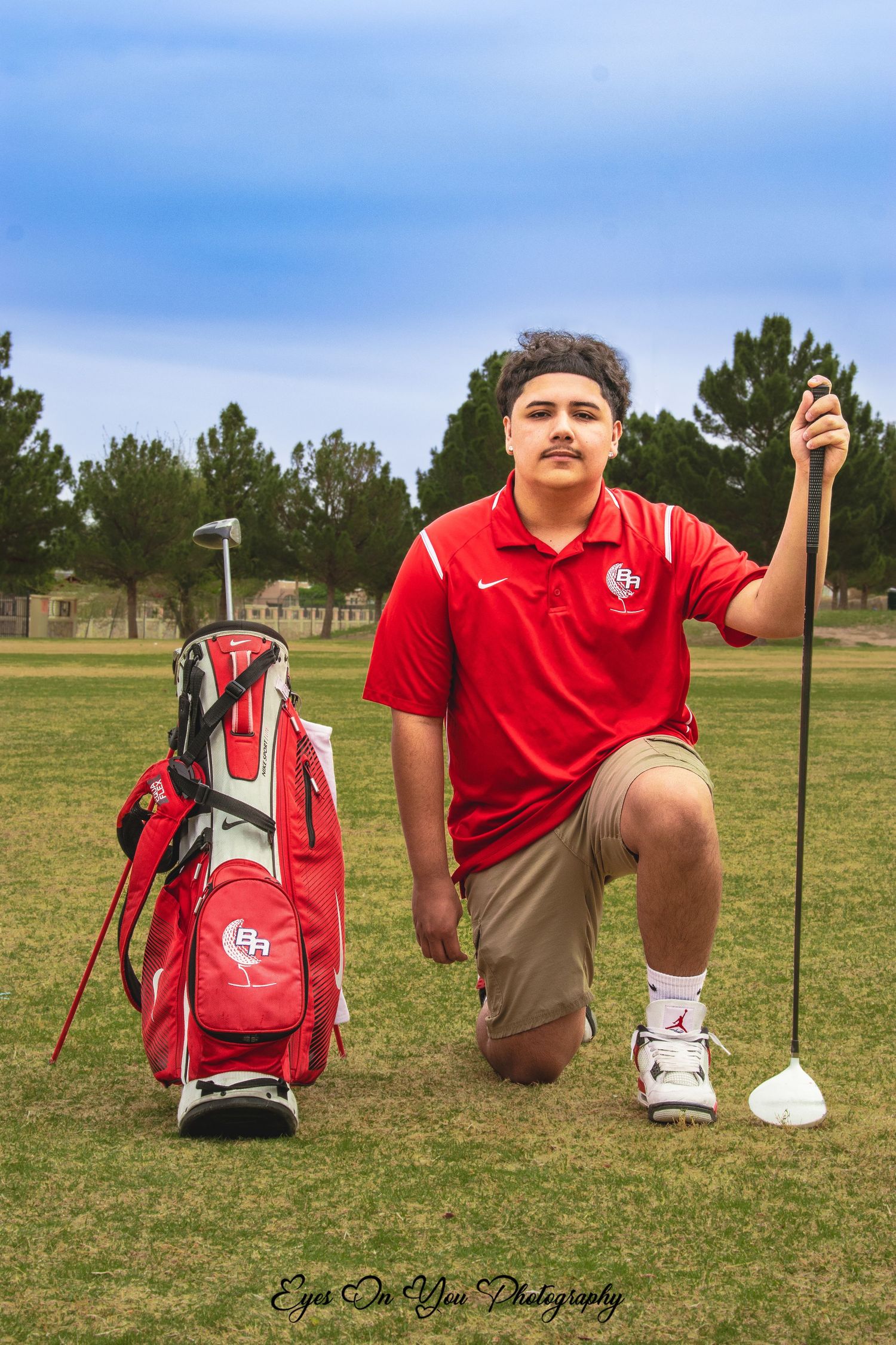 Golfer in red polo poses with club and golf bag on course.