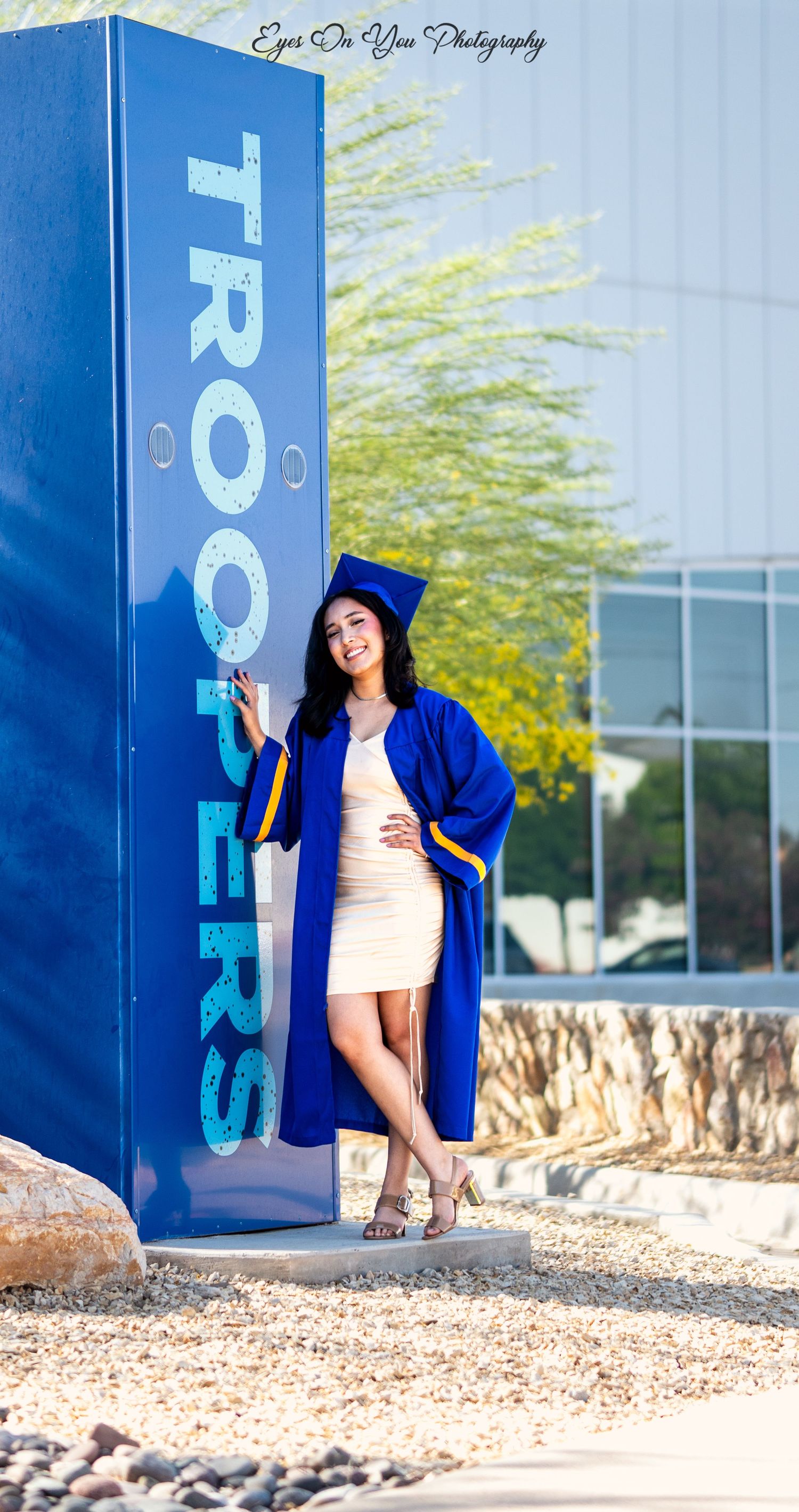 Graduate in blue regalia poses next to large blue Troopers sign on campus.