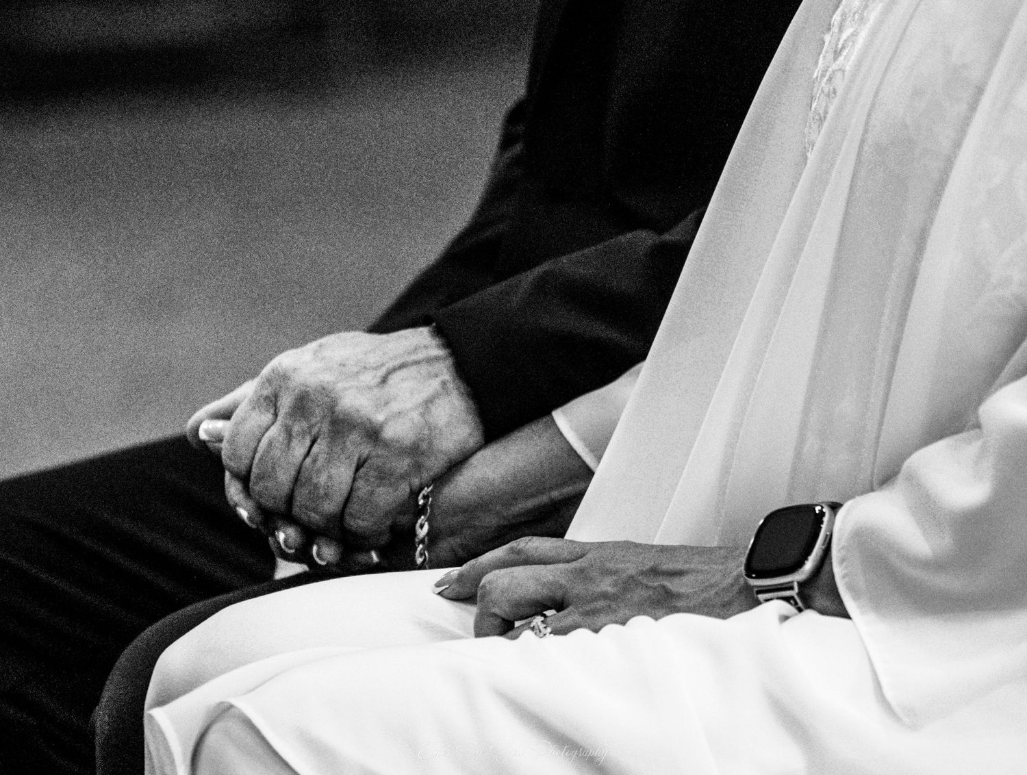 Black and white close-up photo of hands clasped together between figures wearing dark suit and white wedding dress.