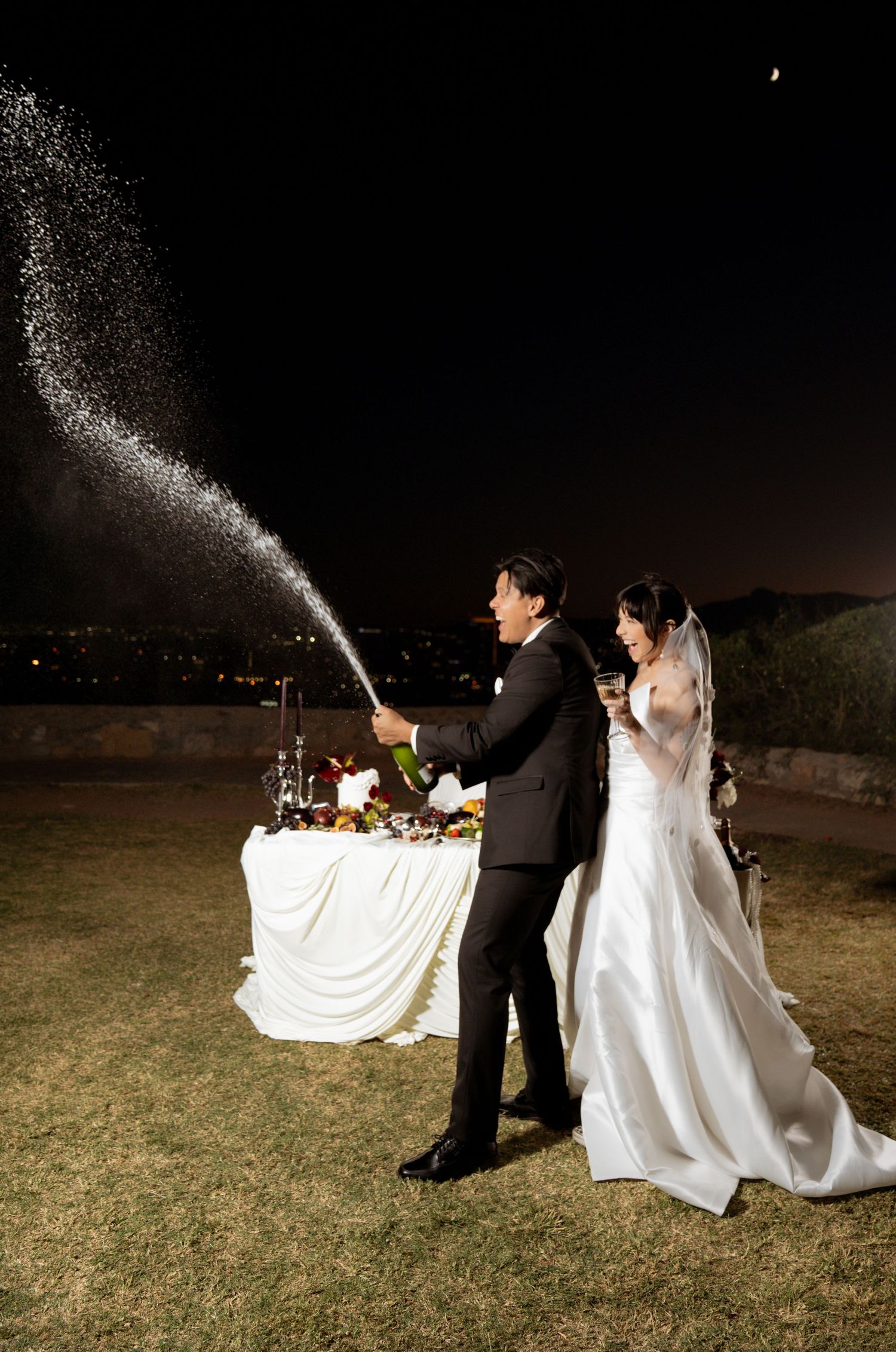 A couple in wedding attire spraying champagne at night in a celebratory moment on a grassy lawn.