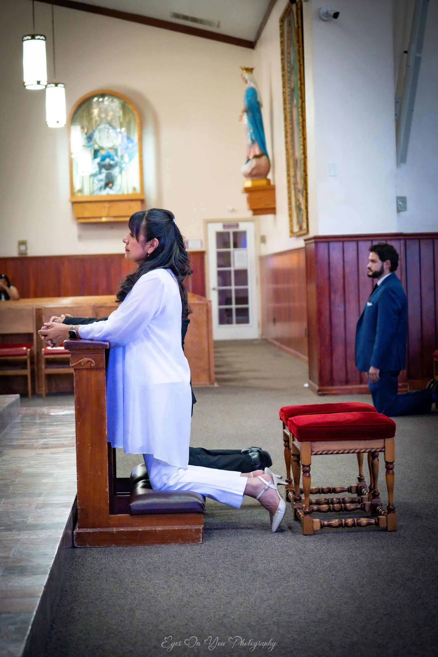 Two figures sit and pray in a church interior with wood paneling and arched windows in a contemplative scene.