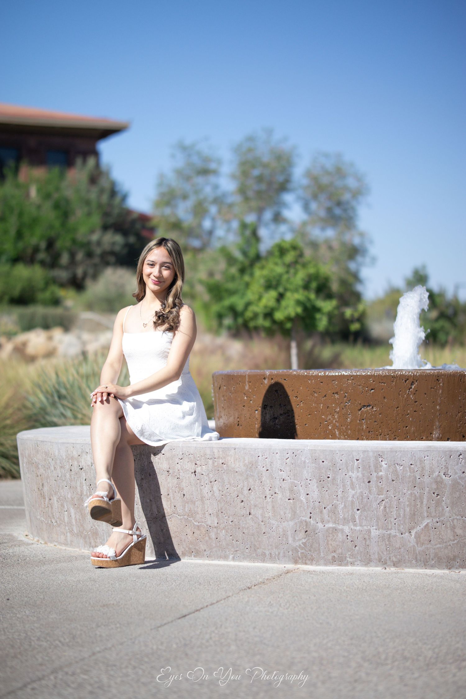 Graduate sitting on concrete ledge near fountain in sunny courtyard setting.