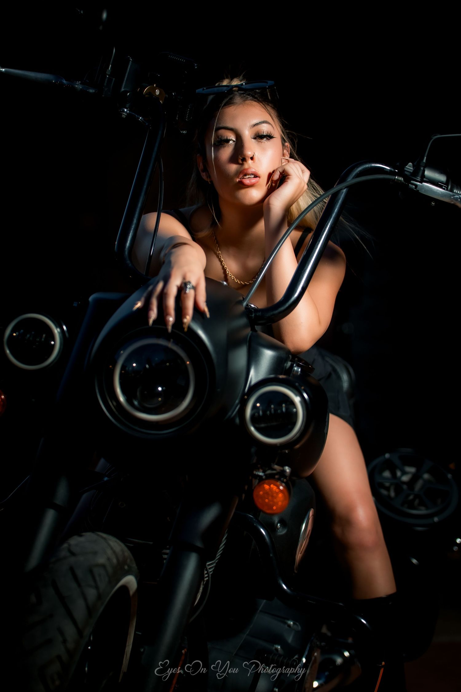 Dramatic low-key portrait of someone posing on a dark motorcycle with dual headlights against black background.
