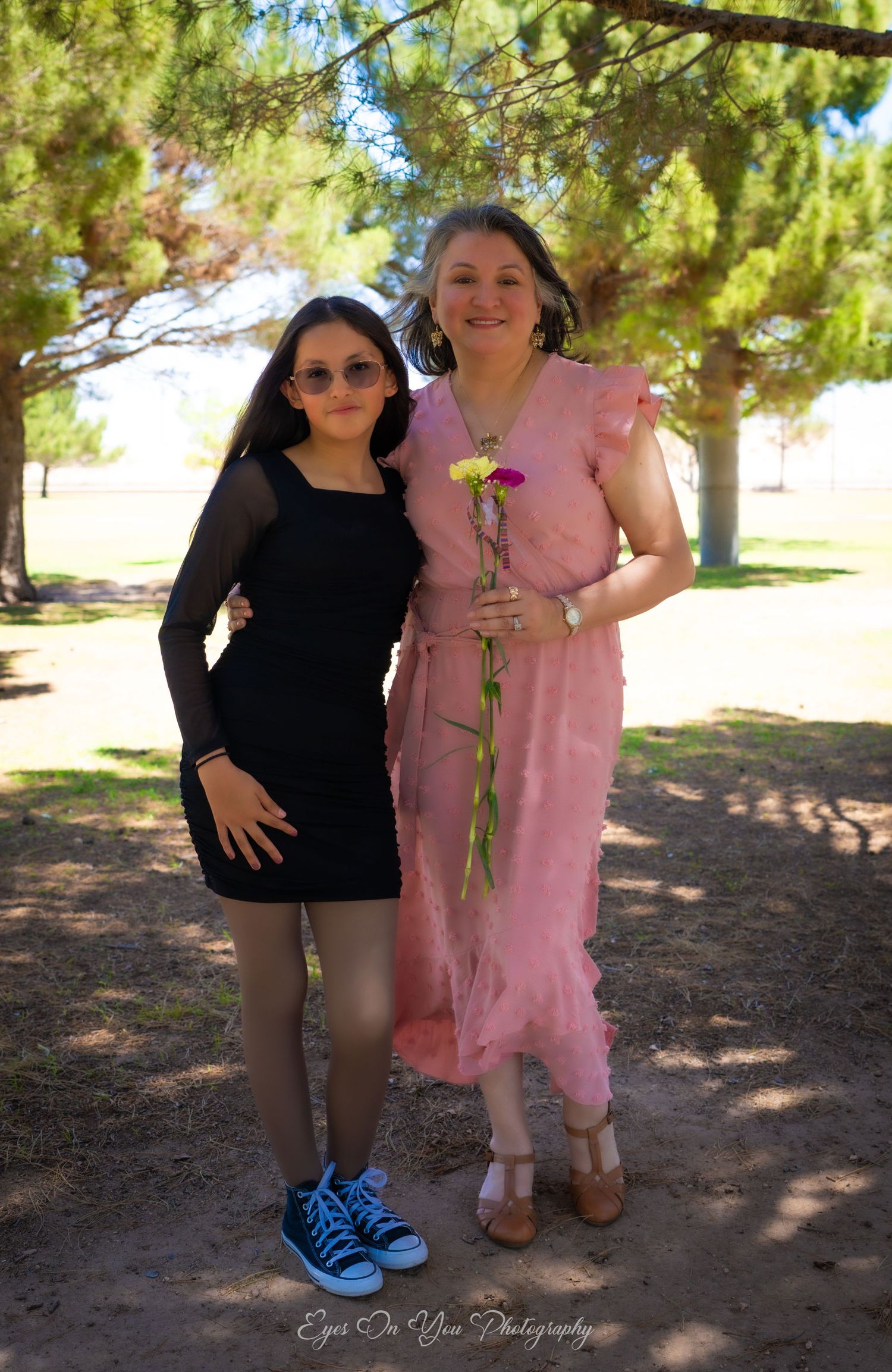 Two people pose outdoors under pine trees, one wearing pink and one in black with bright blue shoes.