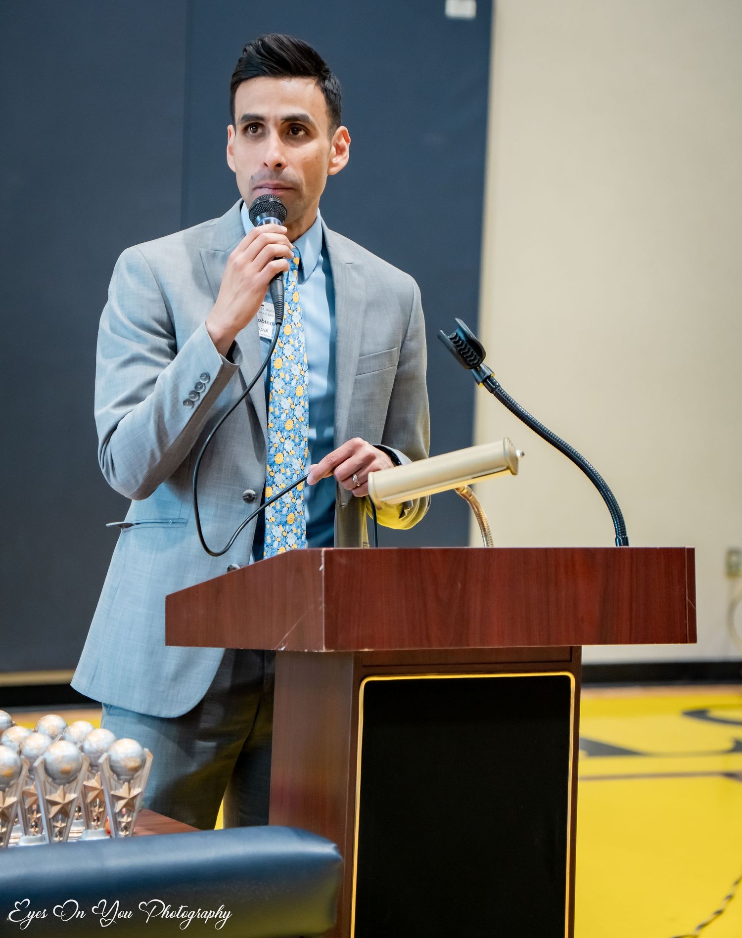 Speaker in light blue suit addresses audience from podium at school event.