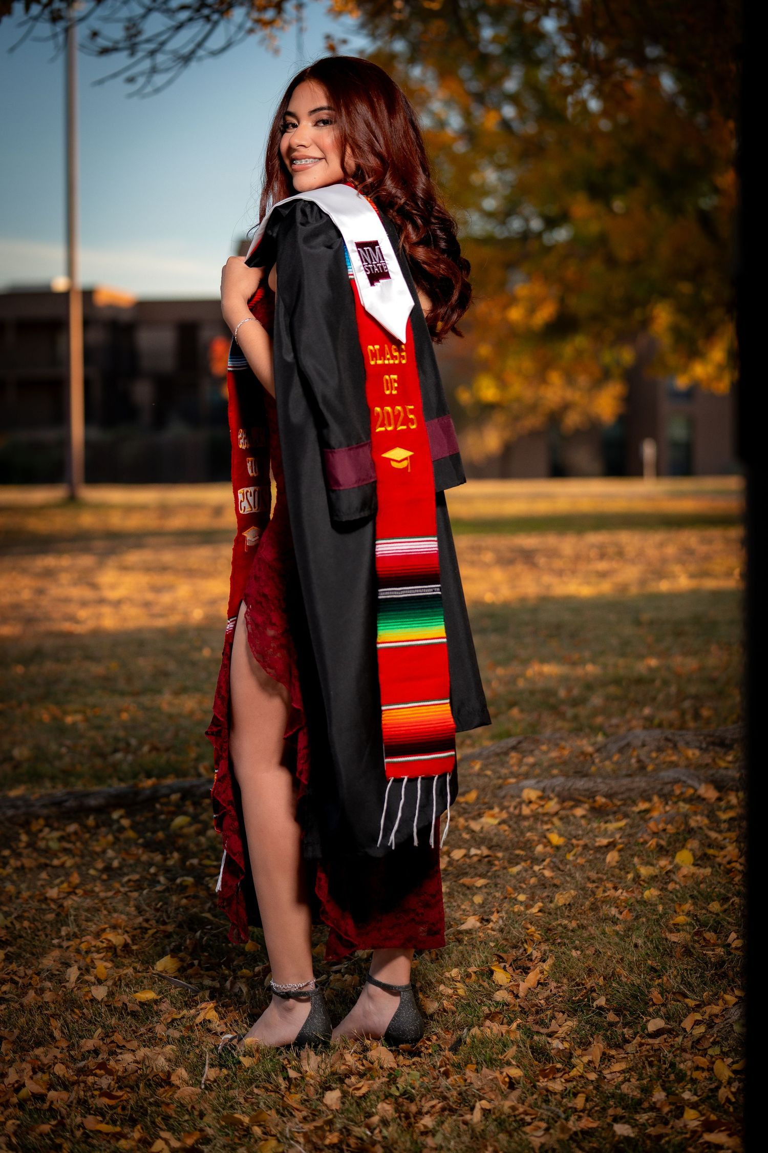 Graduate wearing academic regalia and colorful stole poses in autumn setting with fallen leaves on ground during sunset.