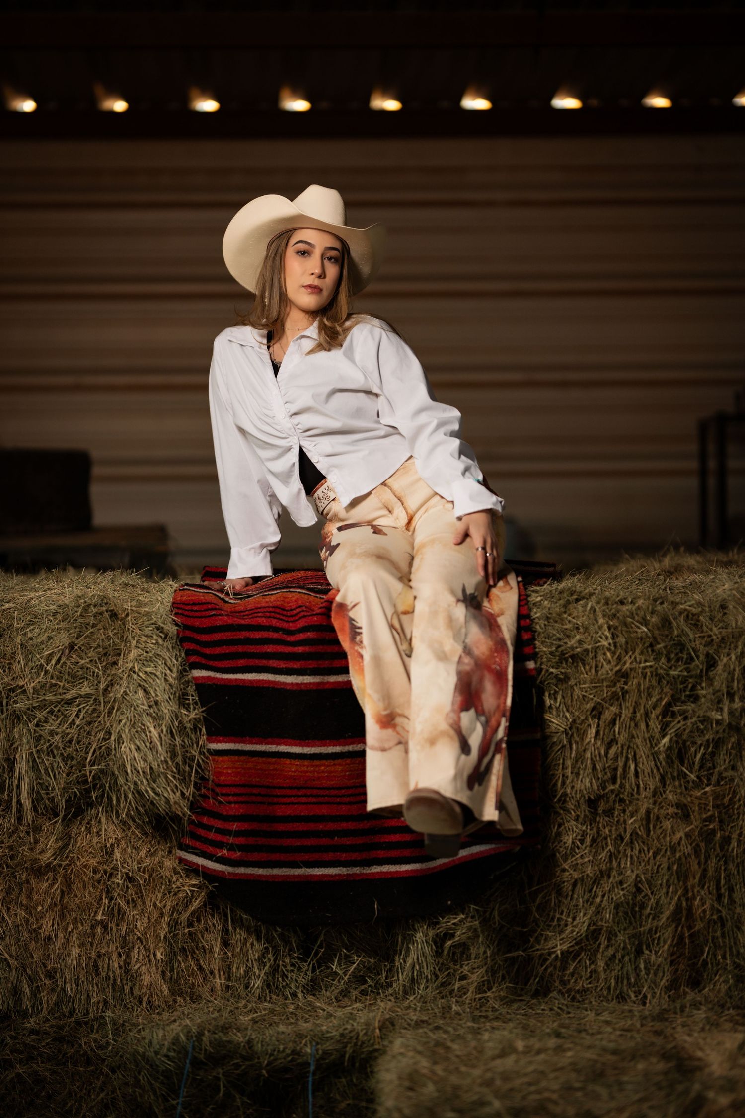 A figure in a white shirt and cowboy hat reclines on hay bales against a dark rustic backdrop.