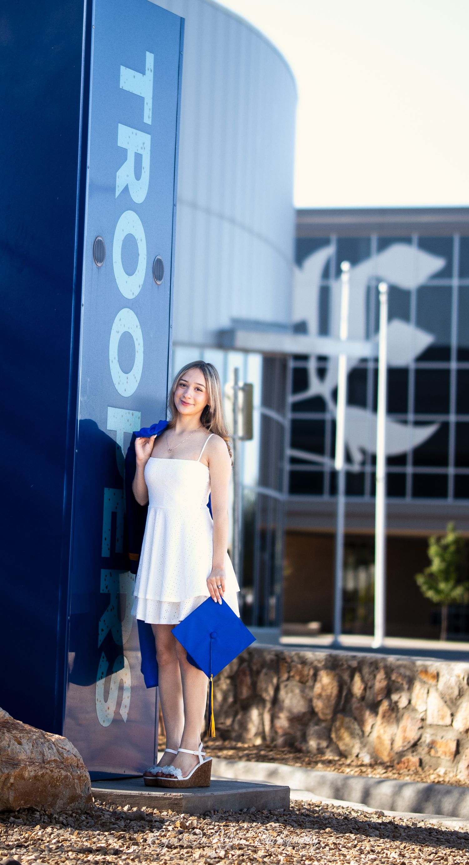 Graduate in white dress holds blue cap near school signage during photo session.