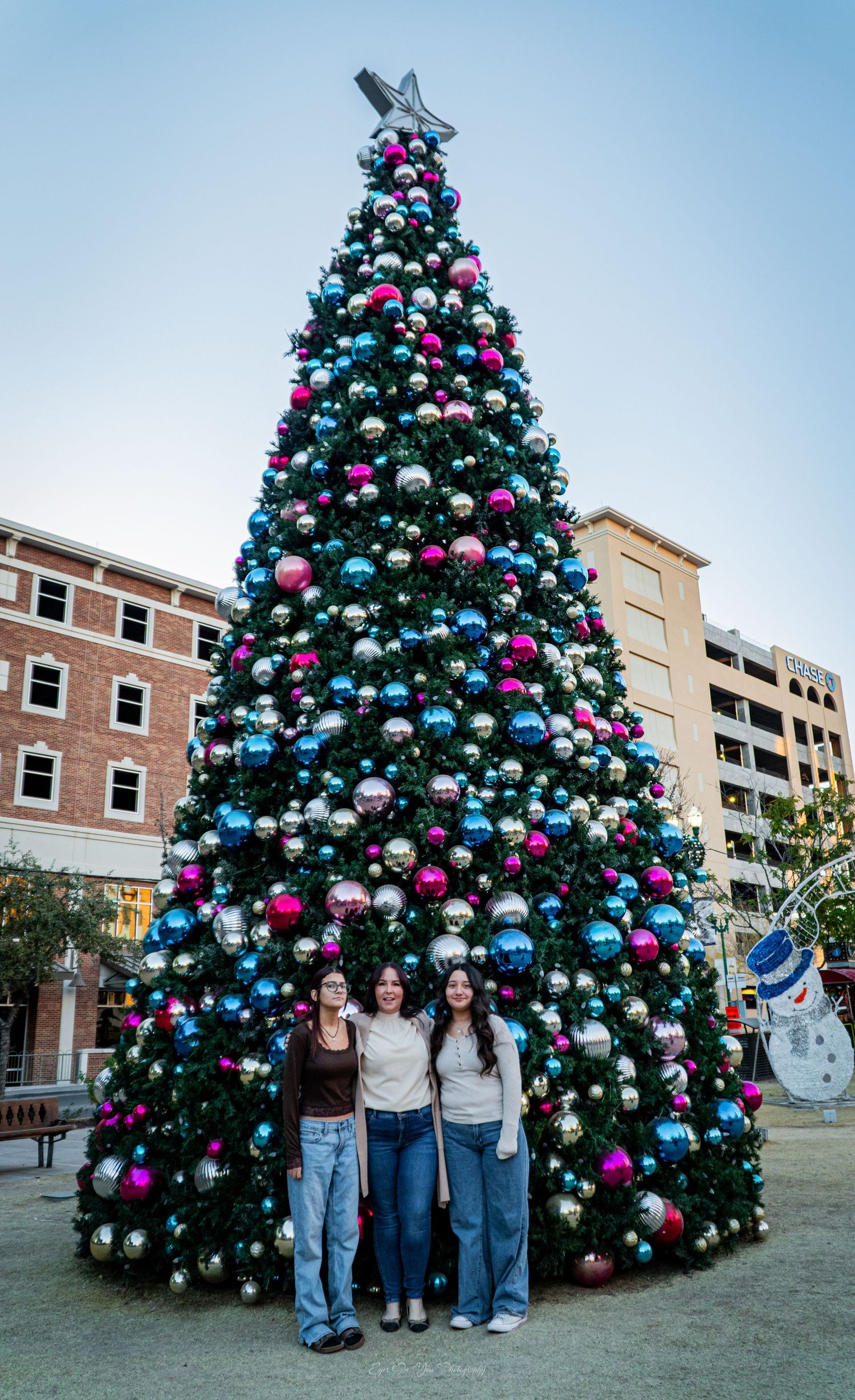 Large decorated Christmas tree with blue and pink ornaments stands in front of brick buildings.