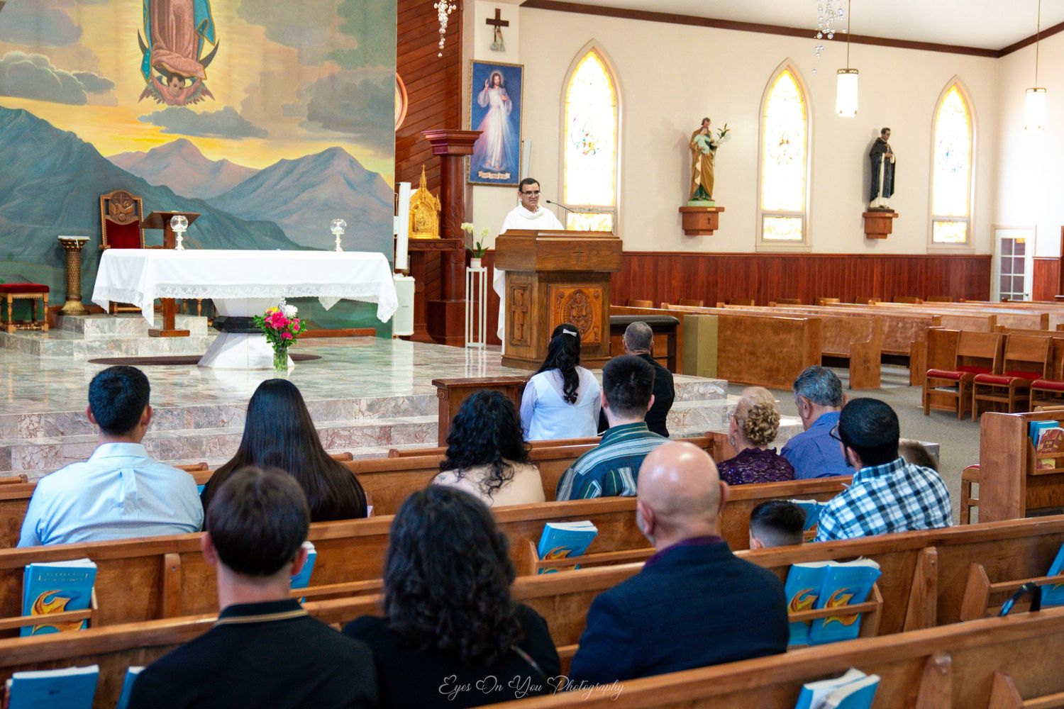 People gathered in wooden pews attend a Catholic church service with religious artwork and statues visible on the walls.