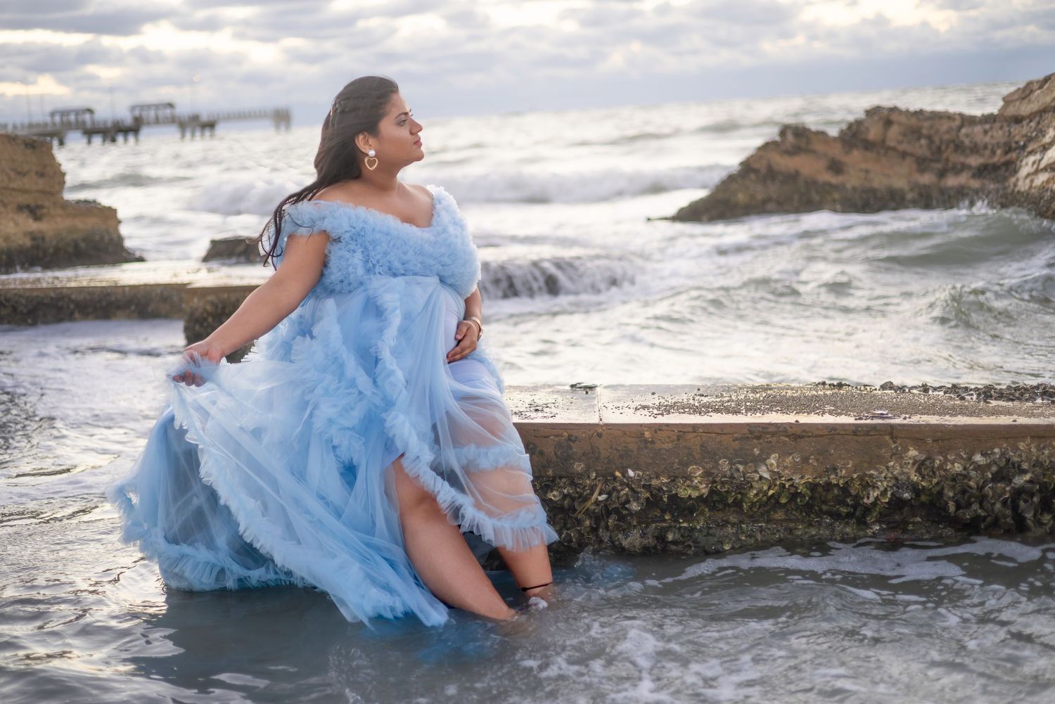 Pregnant woman in blue tulle gown sitting on rocky beach wall with ocean waves, maternity photoshoot.
