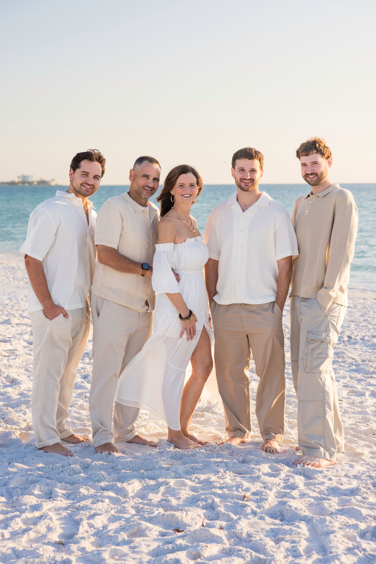 Five adults in coordinated beige and white outfits pose together on a sunny white sand beach with turquoise water.