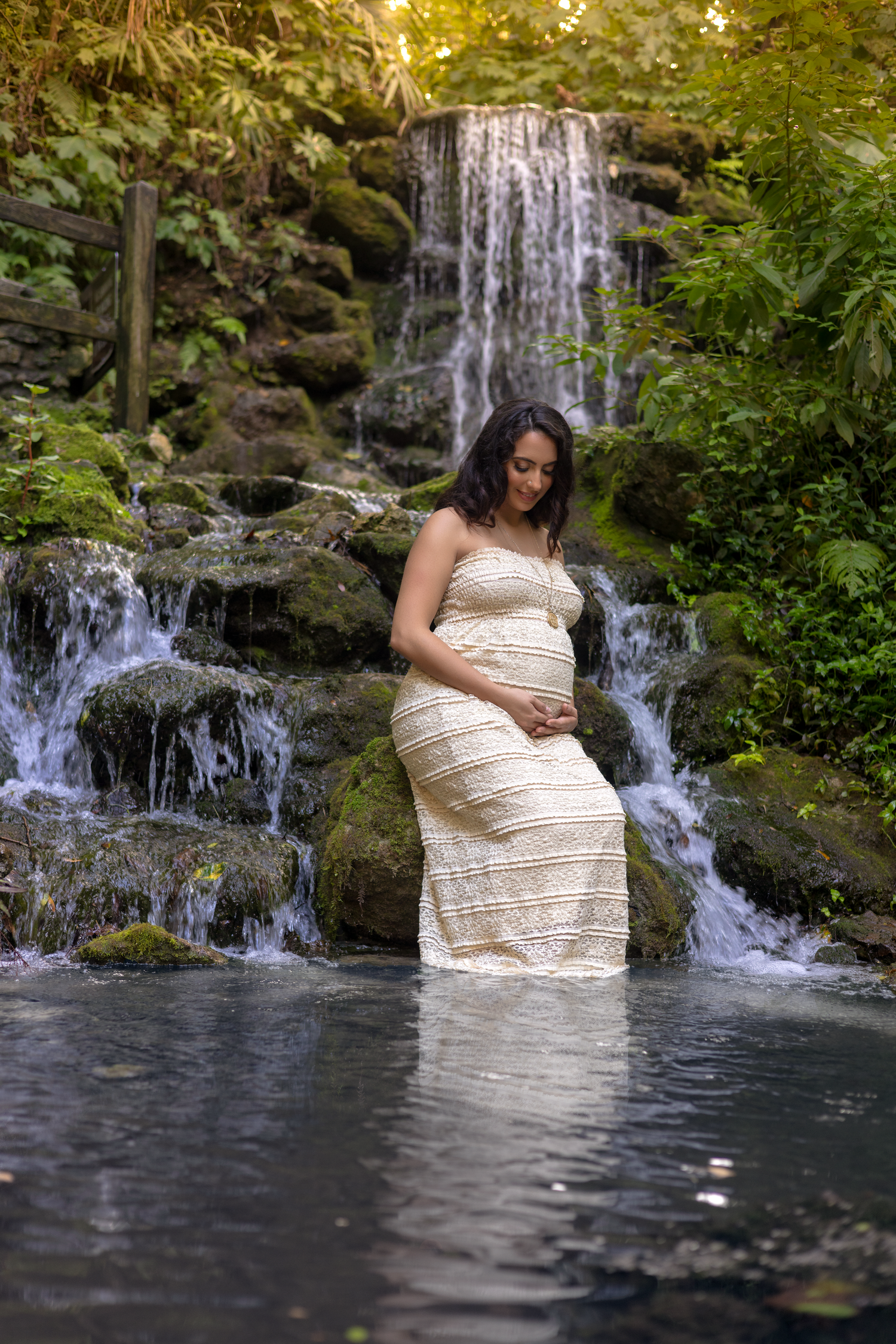 Pregnant woman in white lace dress standing in a serene waterfall pool surrounded by lush green foliage.