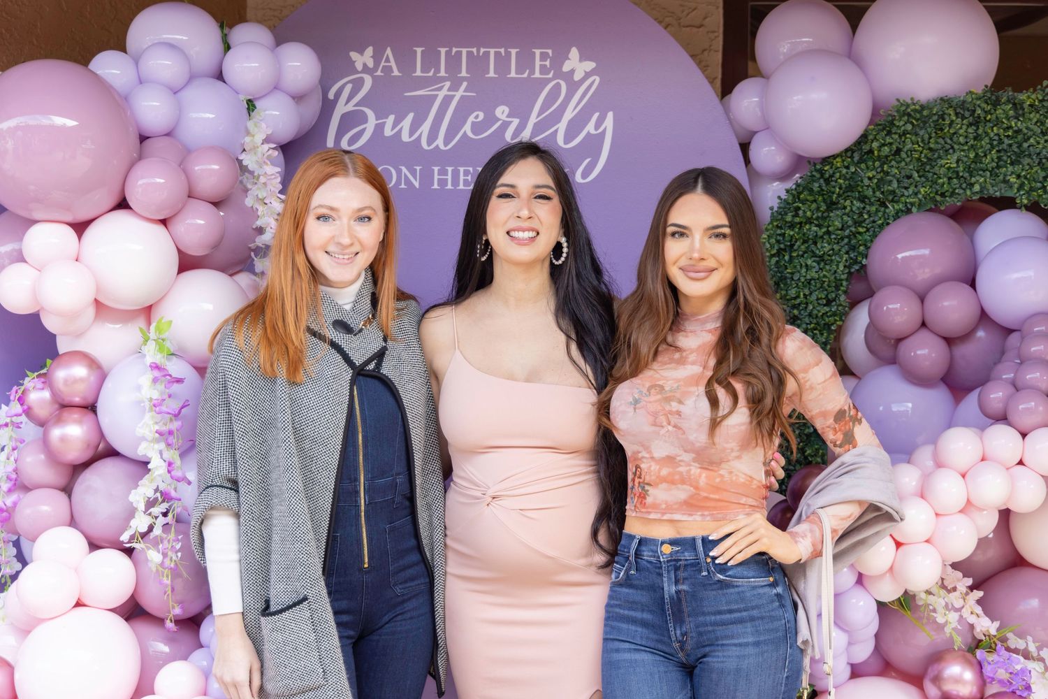 Three women pose at a butterfly-themed baby shower with purple balloon wall backdrop.