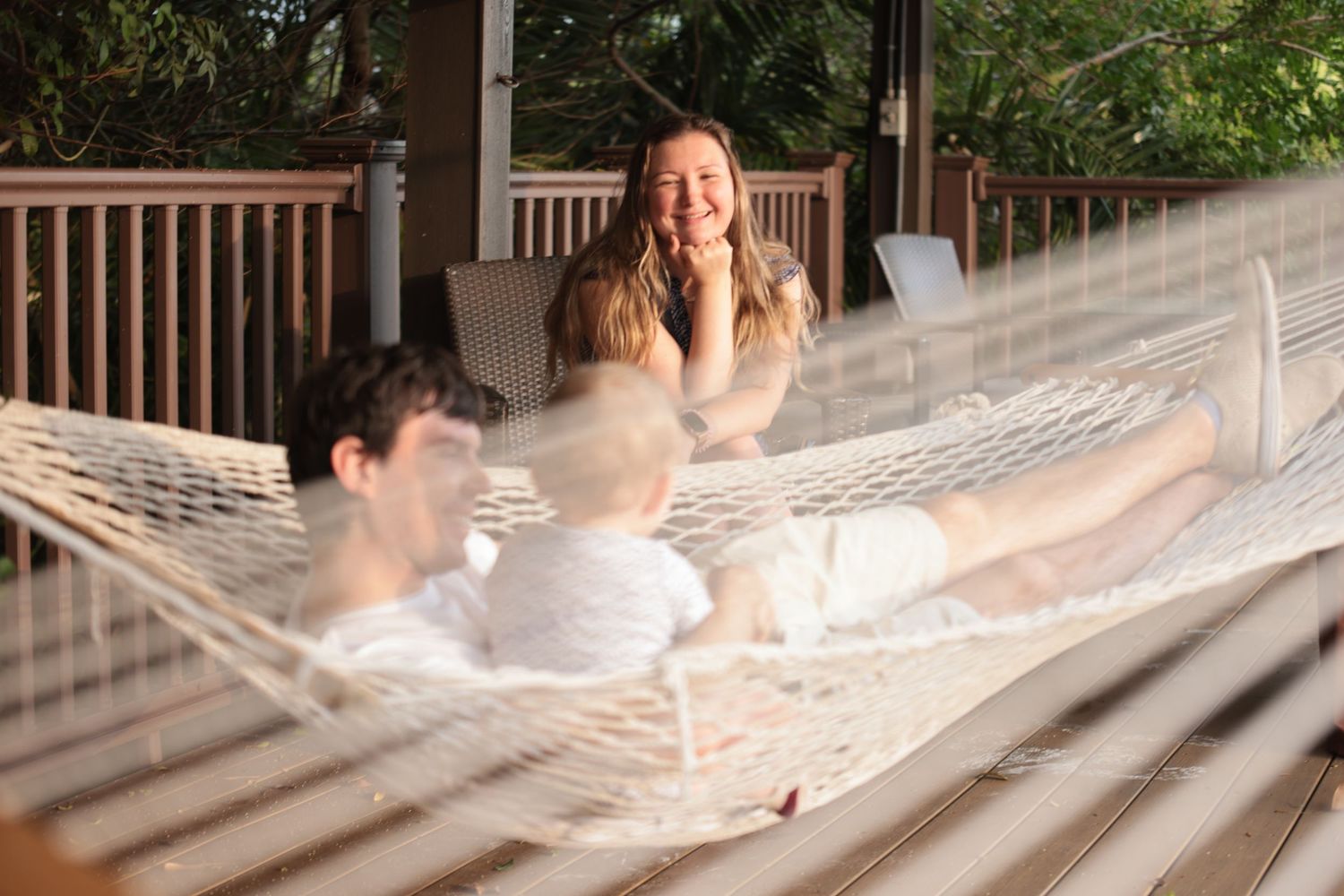 Family relaxing together in a white rope hammock on a wooden deck surrounded by lush green trees.