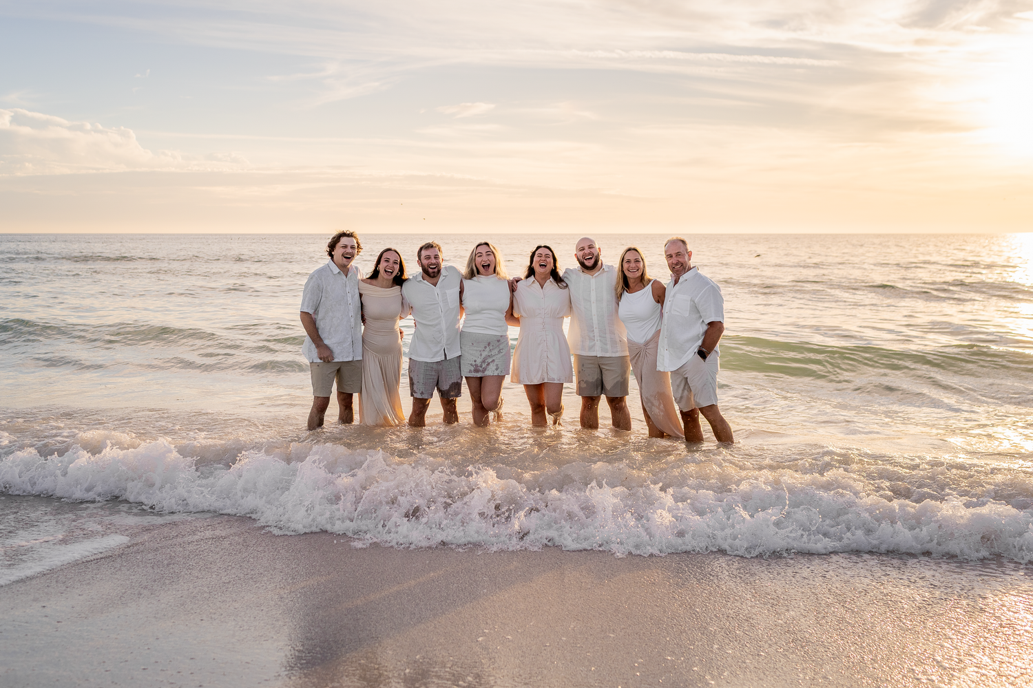 Family in white outfits standing together on a sandy beach at sunset with ocean waves.
