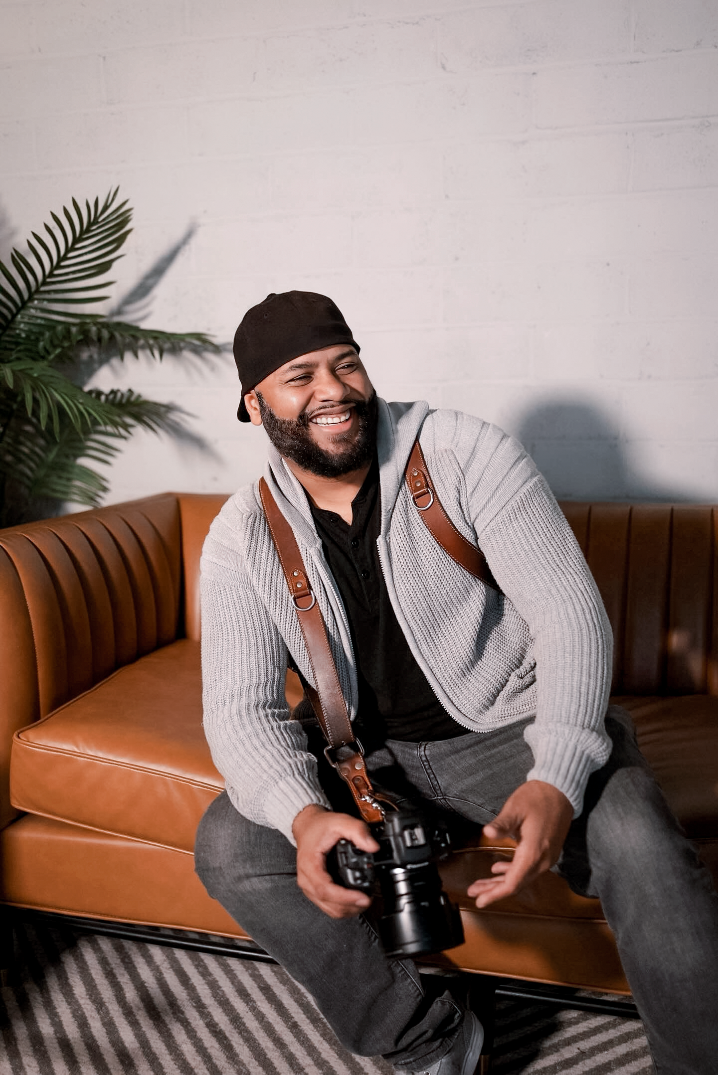 Photographer with camera sits on brown leather couch wearing gray cardigan and beanie while smiling warmly.