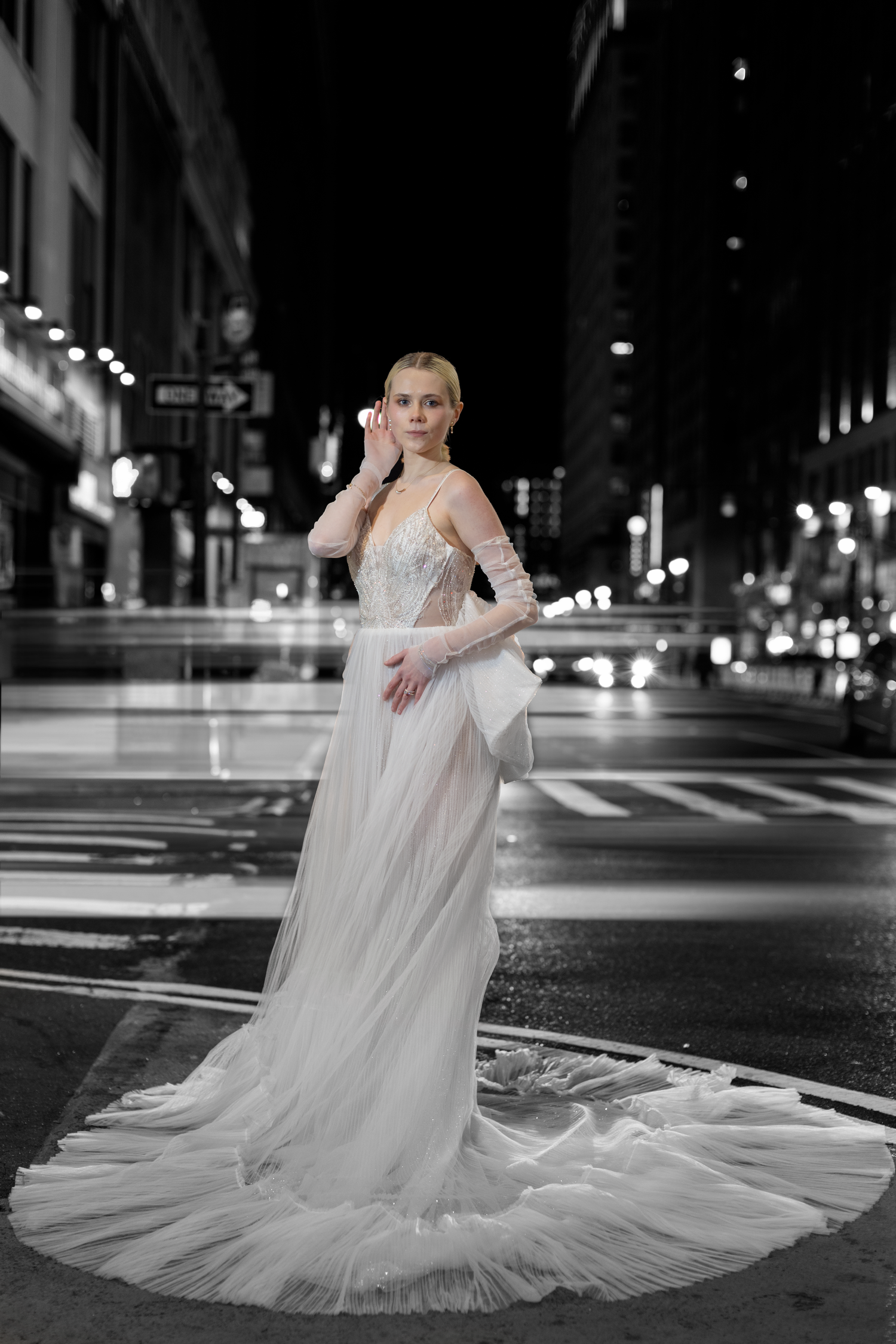 A dramatic bridal portrait in a flowing white gown standing on a city street at night with twinkling lights in the background.
