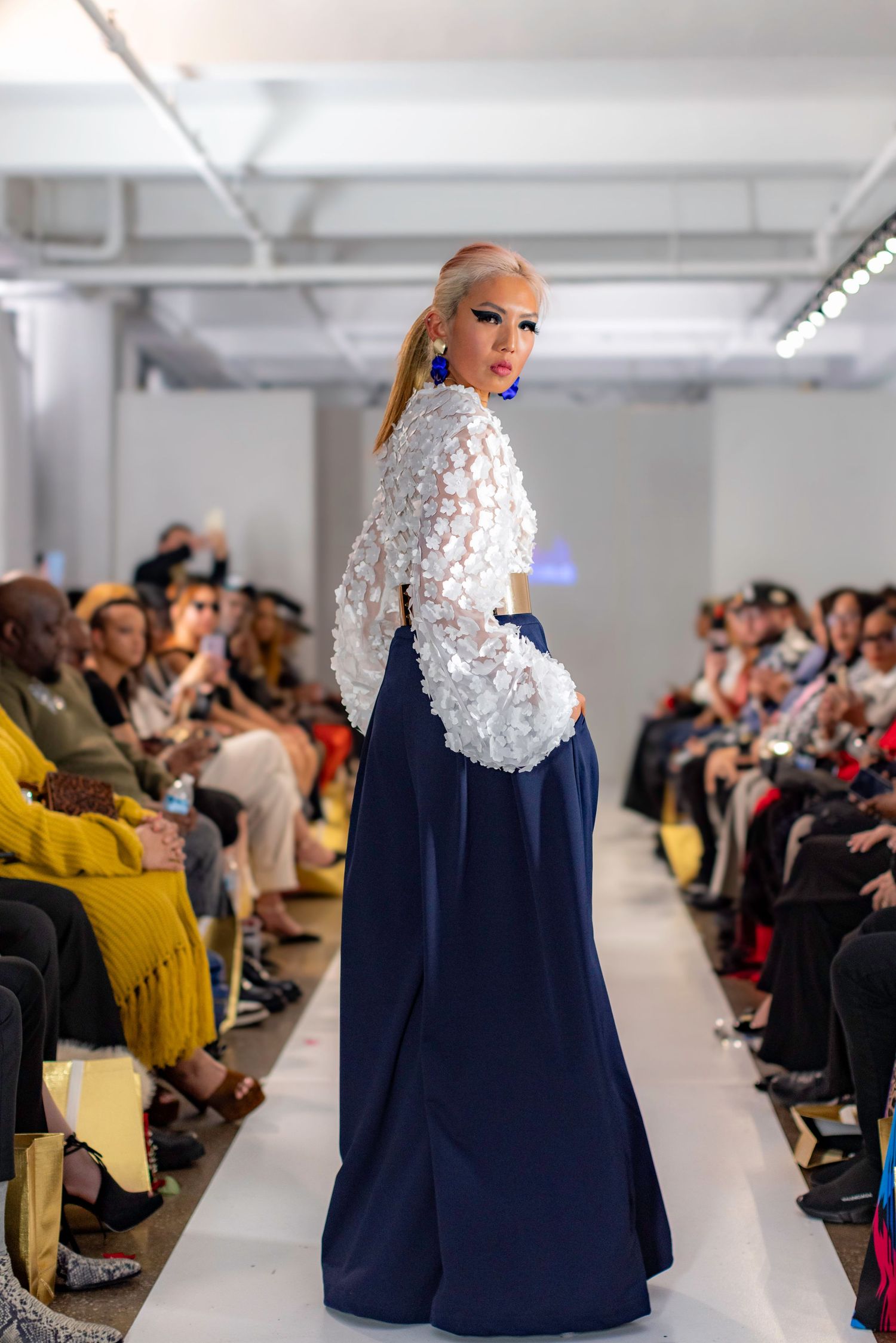 A model walks the runway wearing an elegant white lace top with navy blue ball gown skirt during a fashion show.