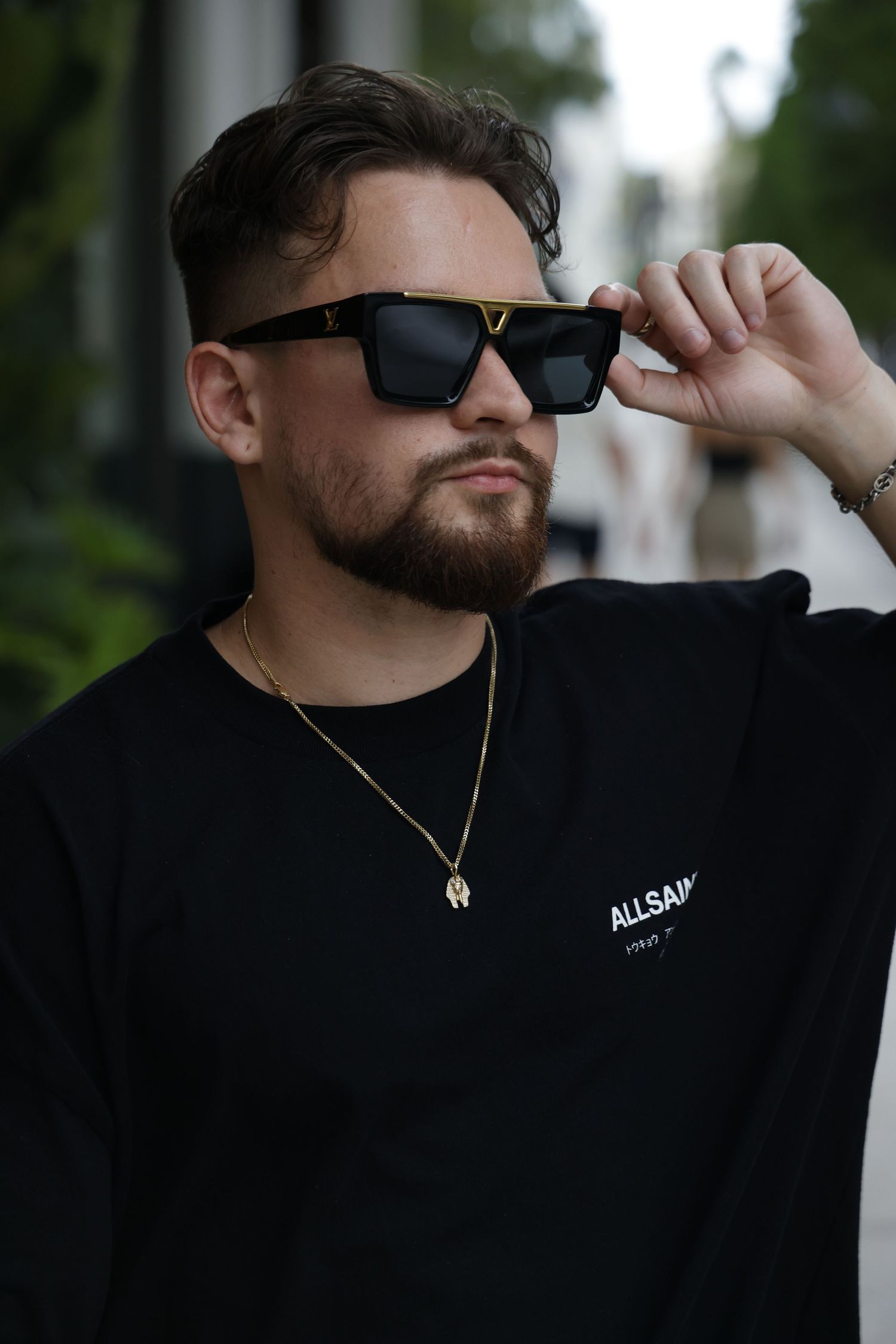Stylish portrait of someone wearing black sunglasses, gold necklace and AllSaints black sweatshirt in outdoor setting.