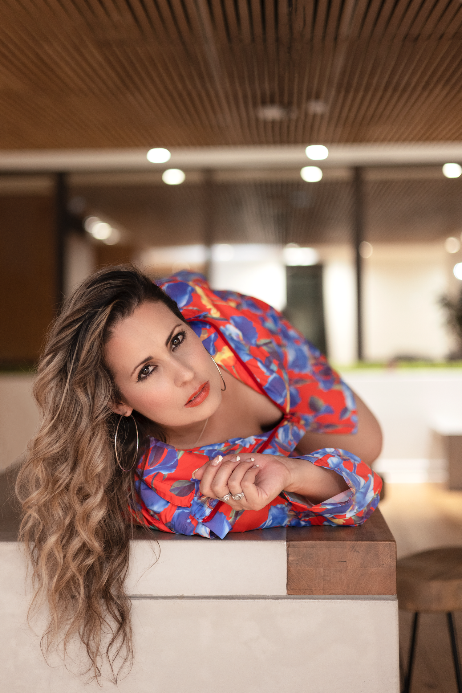 A person wearing a vibrant red and blue floral dress poses elegantly while leaning on a surface in an indoor setting.