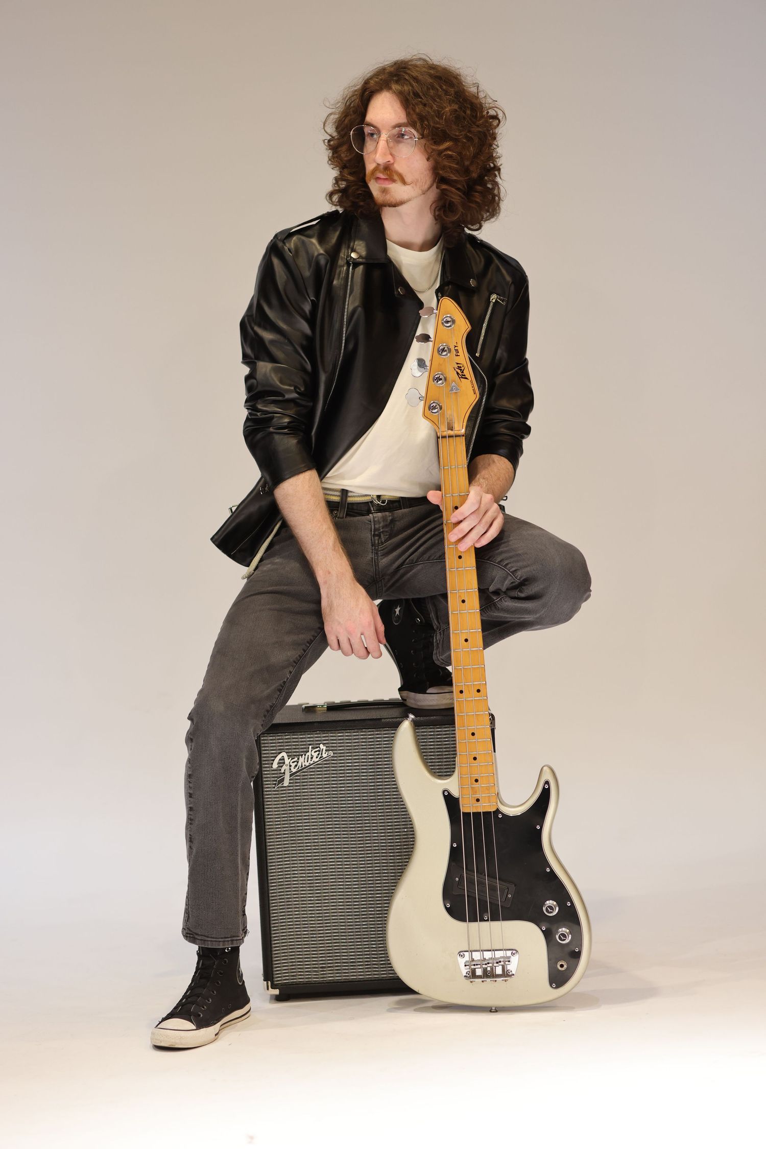 Musician in leather jacket sits on Fender amp holding white Precision Bass guitar against studio backdrop.