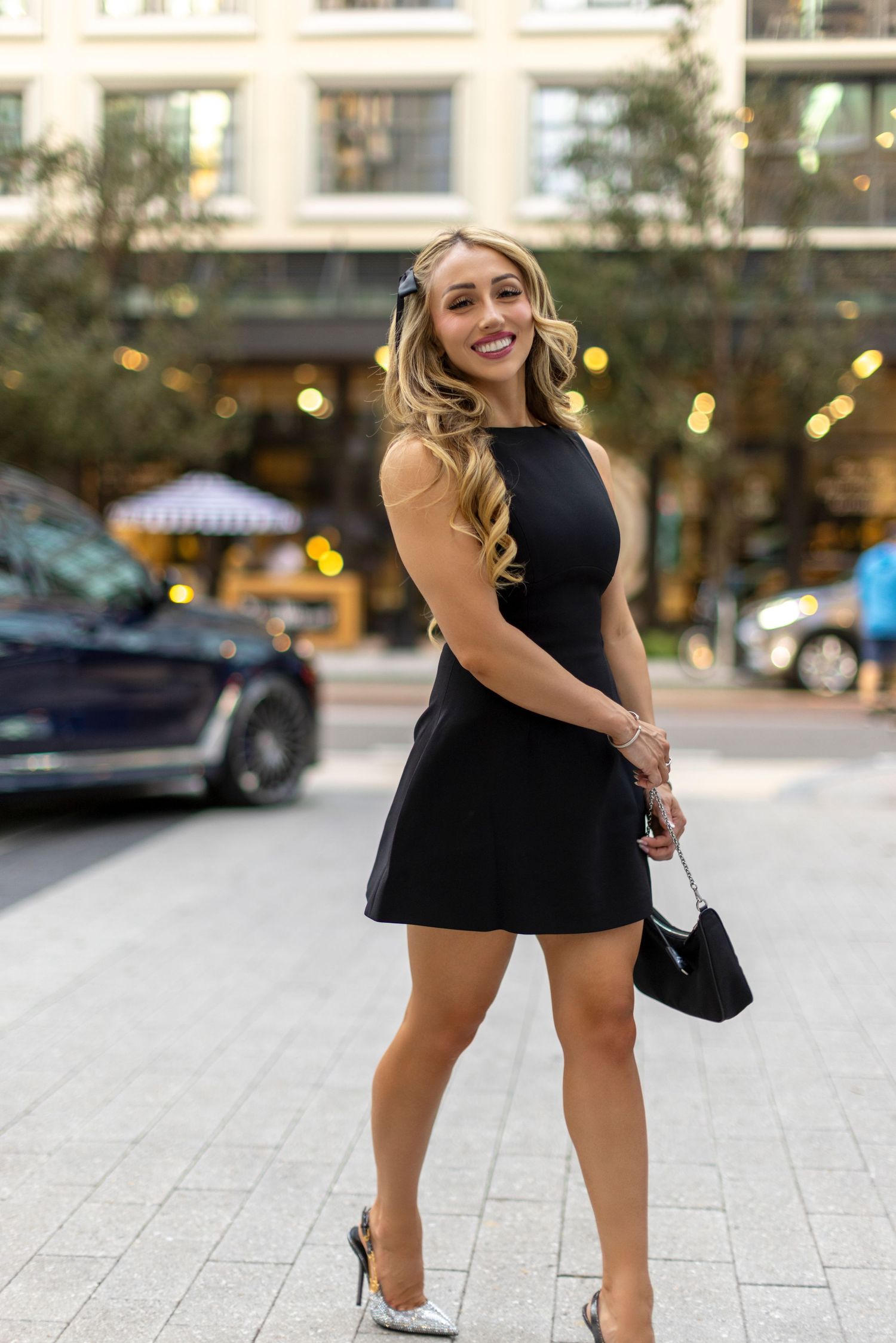A woman in a black sleeveless dress and heels smiles while posing on a city sidewalk near storefronts at dusk.