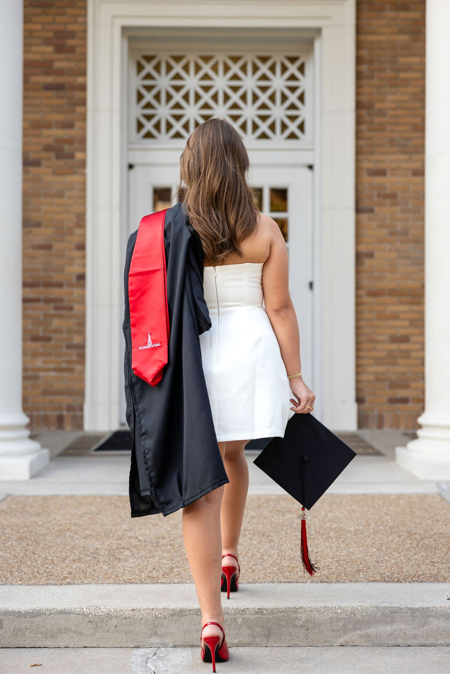 Graduate in white dress holding cap and black gown with red stole, walking toward building entrance in red heels.