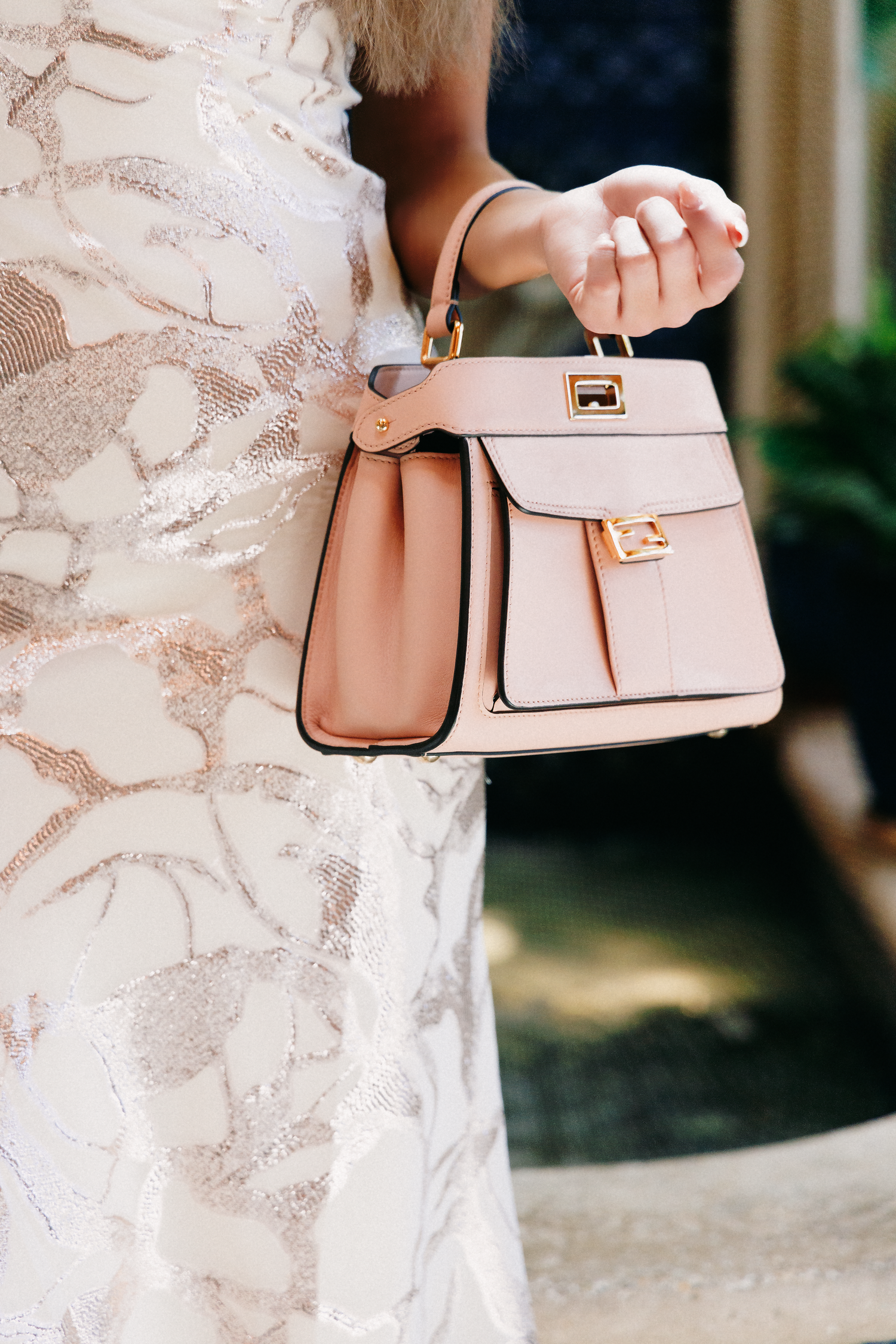 Woman in white lace dress holds a blush pink structured handbag with gold hardware details.