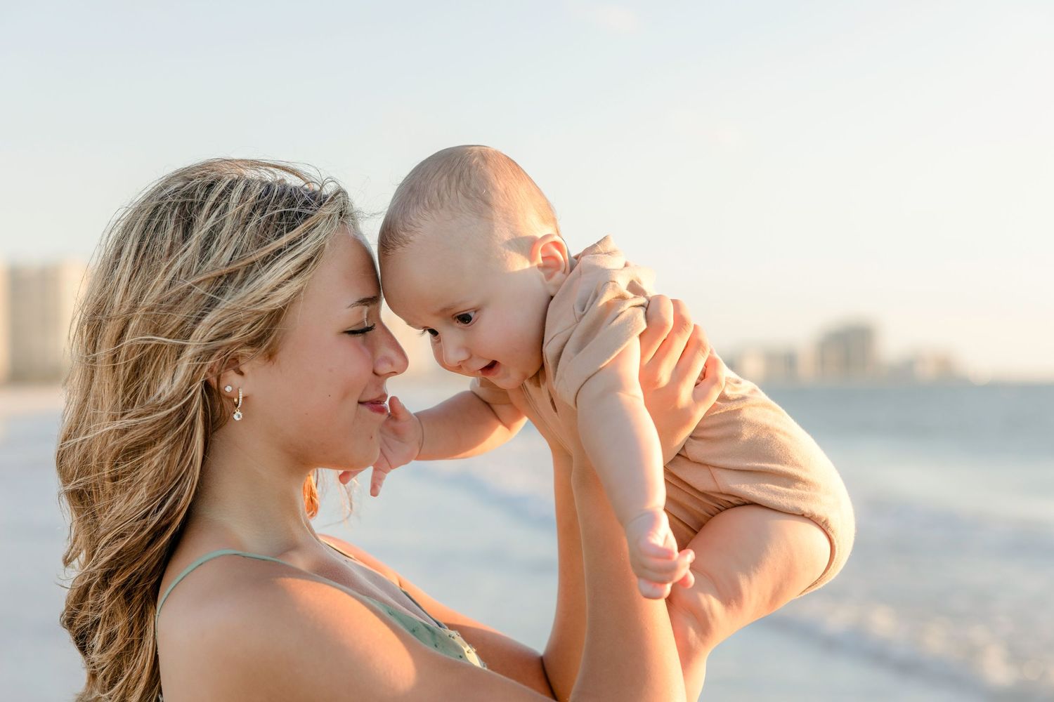 A mother lovingly holds her baby up to her face on a sunny beach, sharing a tender forehead-to-forehead moment.