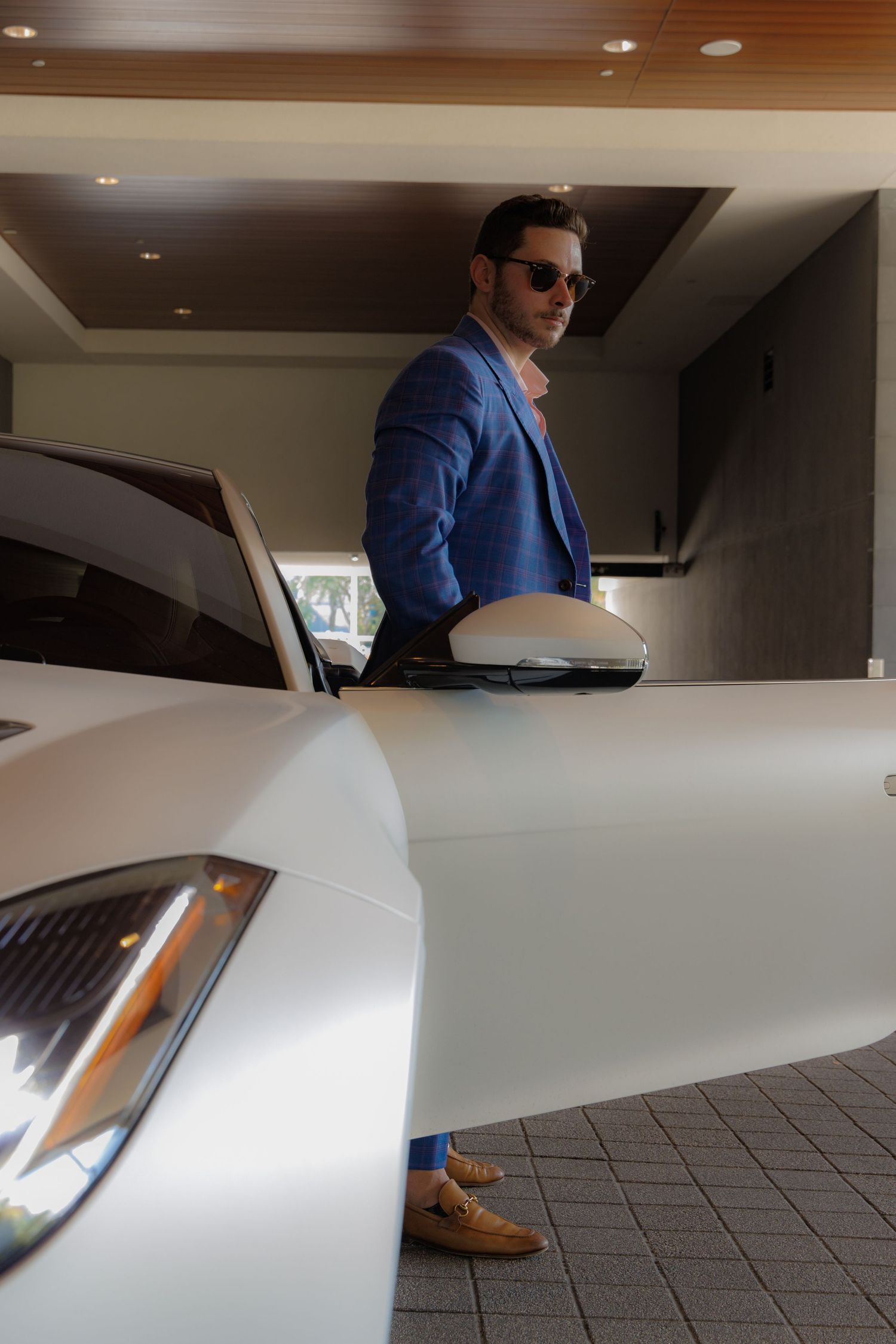 A person in a blue blazer and sunglasses stands next to a white luxury car in a covered parking area.