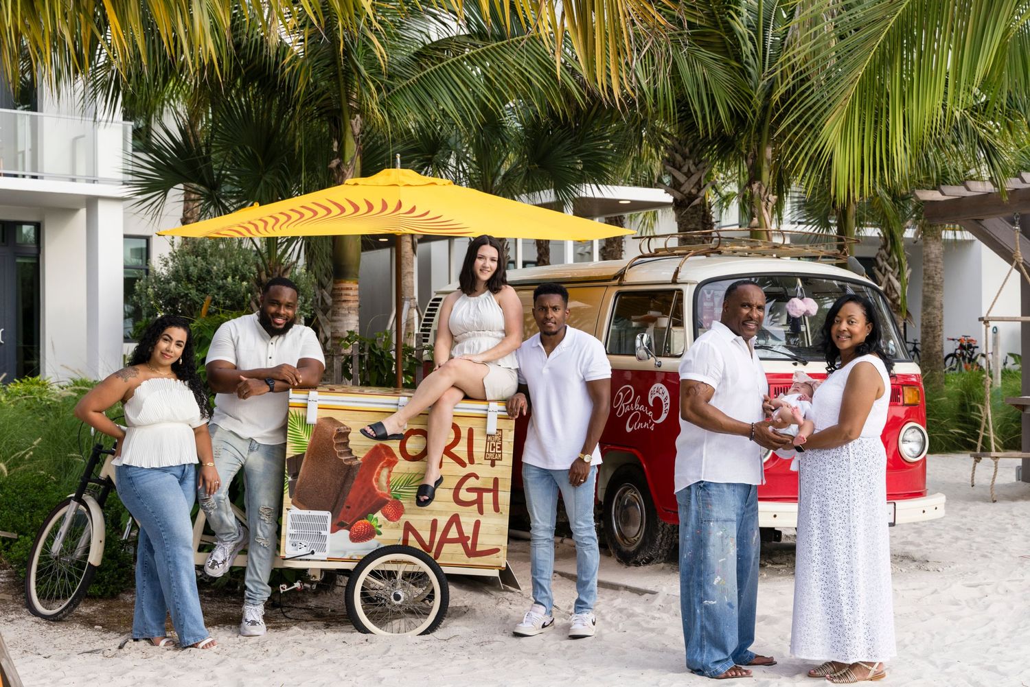 Group of friends posing near a colorful vintage VW bus food cart under palm trees at a tropical resort.