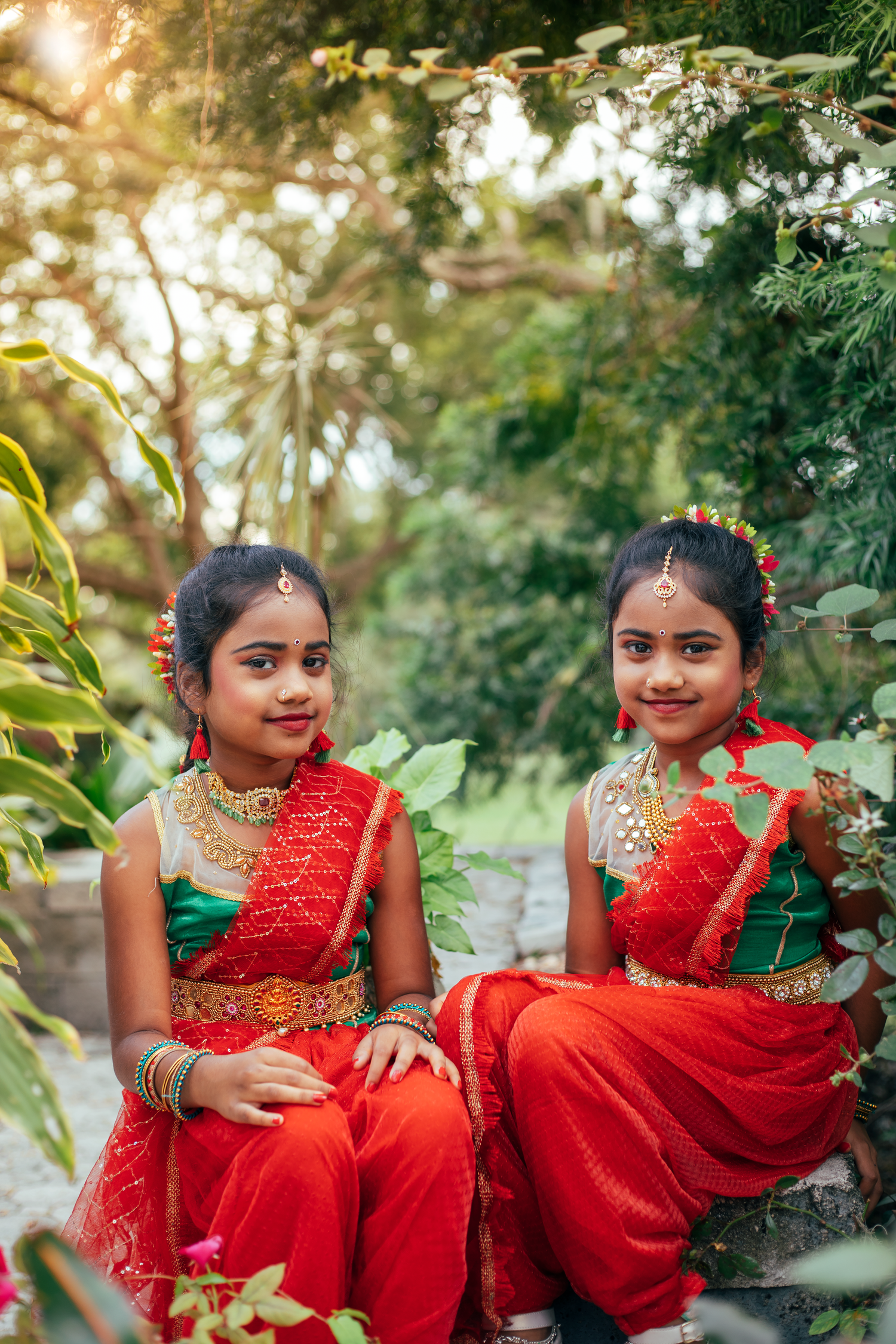 Two young girls in red and green traditional Indian attire with gold jewelry pose outdoors among lush green foliage.