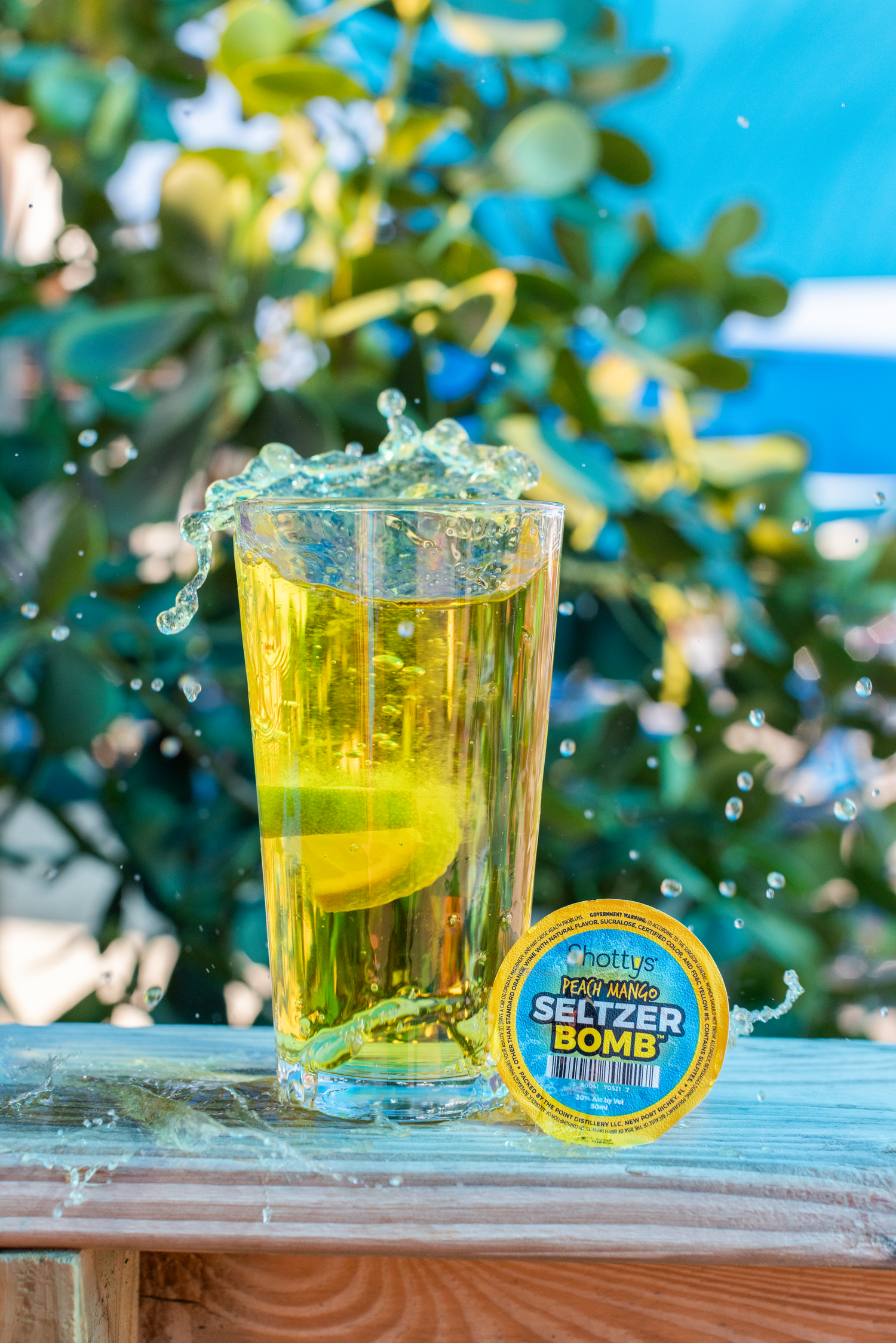 A glass of sparkling beverage with a lemon slice splashes next to a Seltzer Bomb tin on a wooden surface outdoors.