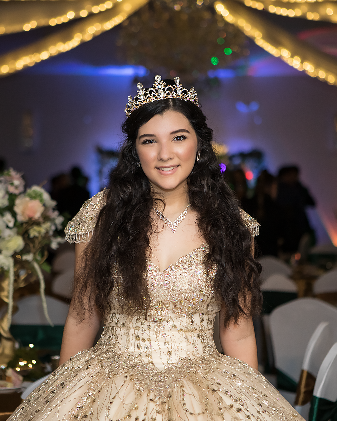 Young woman in gold quinceañera gown and crystal tiara smiles at decorated celebration venue.