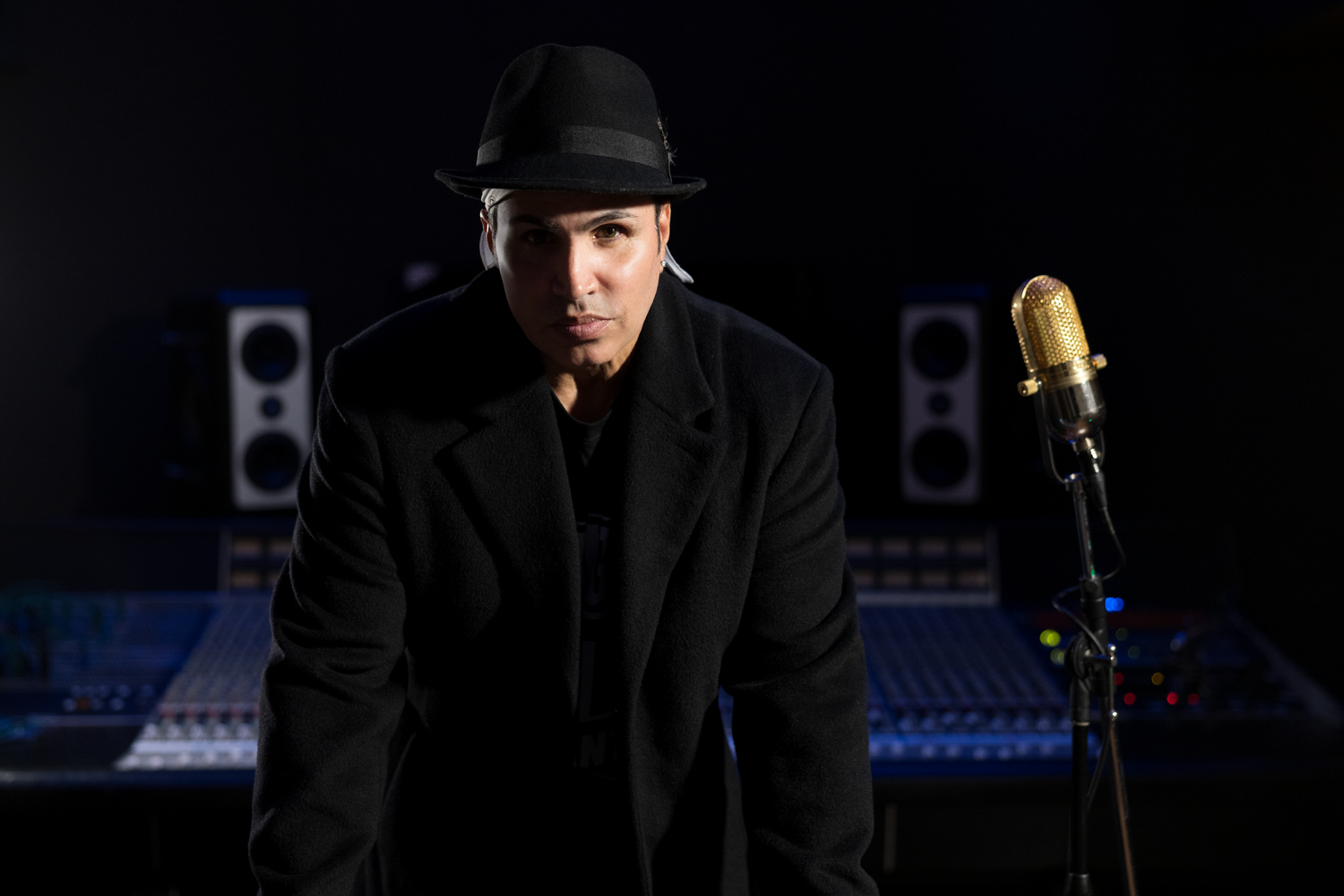 A performer in dark clothes and a black hat stands on stage next to a vintage gold microphone in a dimly lit studio.