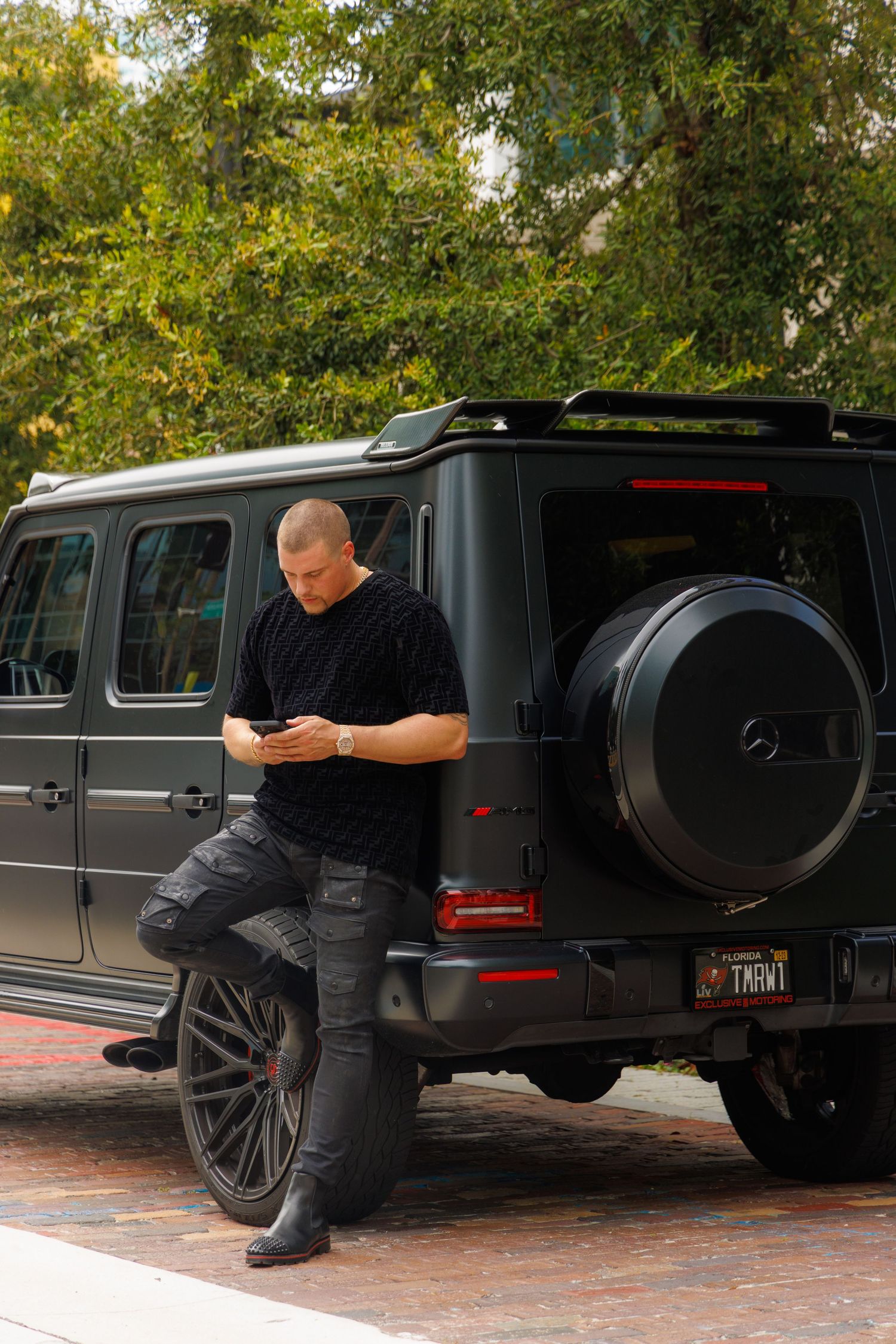 A person in black clothing checks their phone while leaning against a gray Mercedes G-Wagon on a brick driveway.