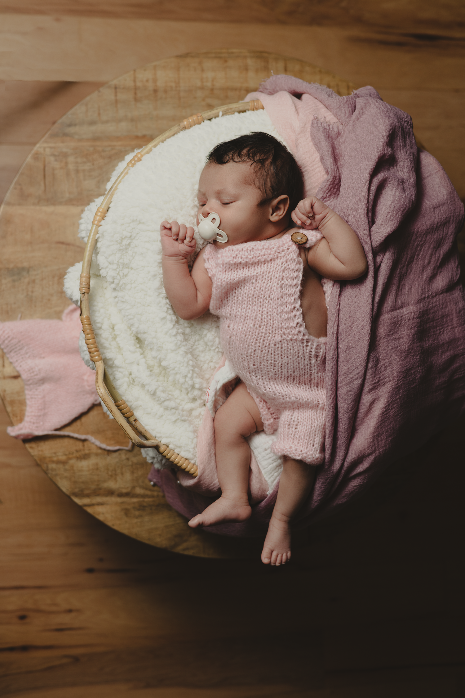 Sleeping newborn baby in pink knit romper with pacifier, nestled in a round basket with soft pink blankets.