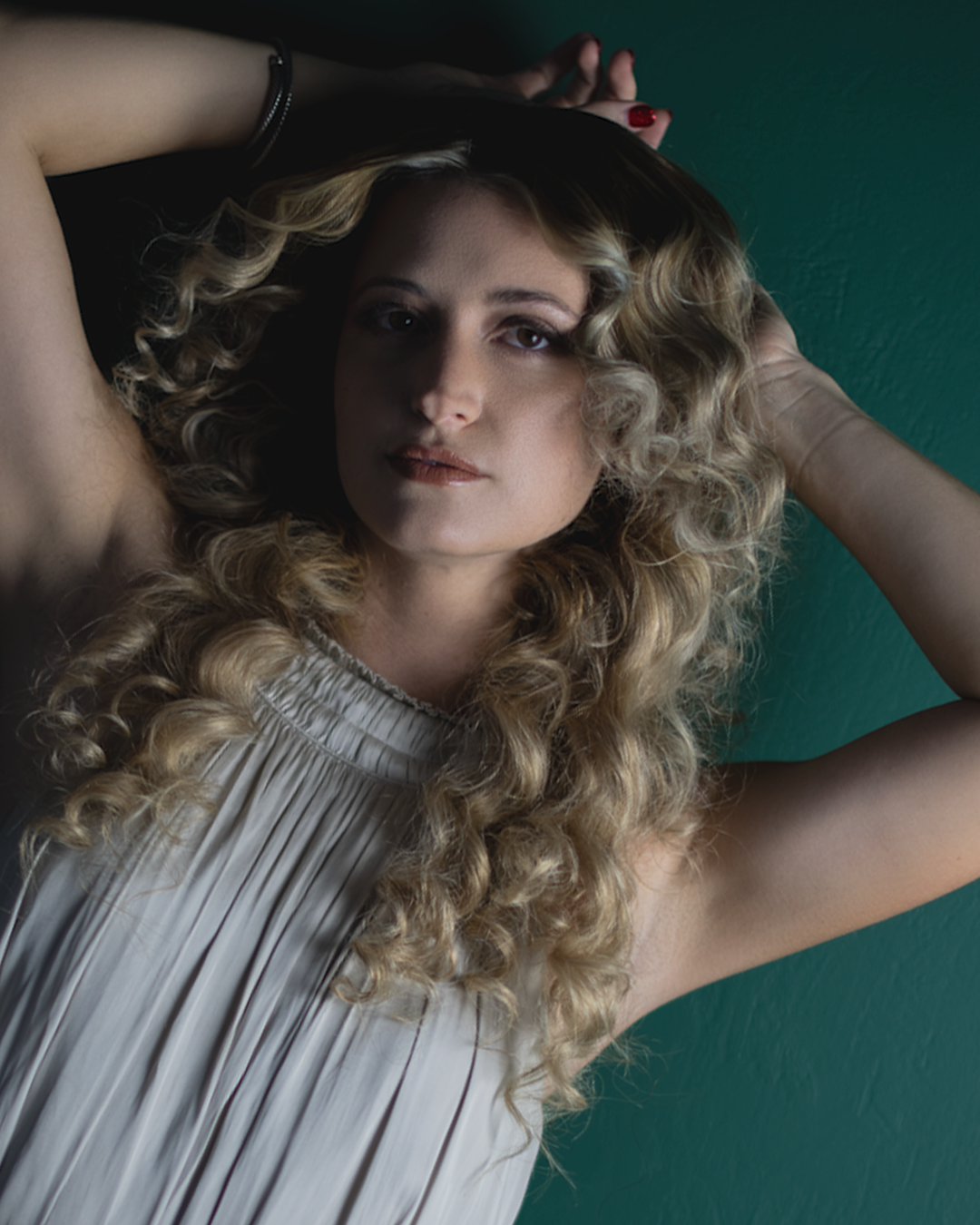 A figure with long curly blonde hair poses dramatically against a teal background in a flowy gray top.