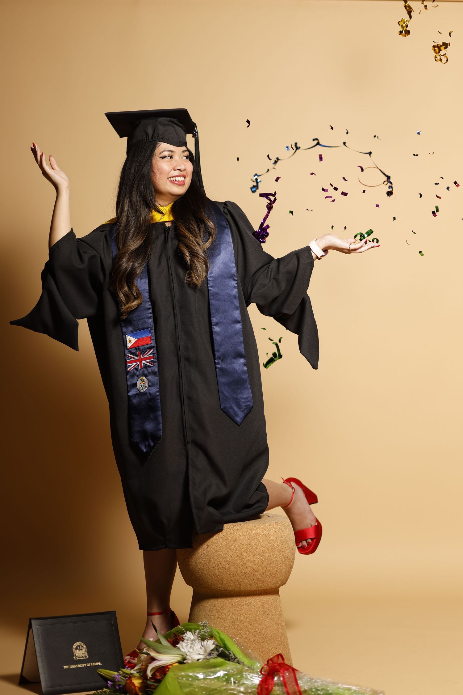 A young woman in black graduation cap and gown celebrates with confetti, wearing red heels beside a diploma and flowers.