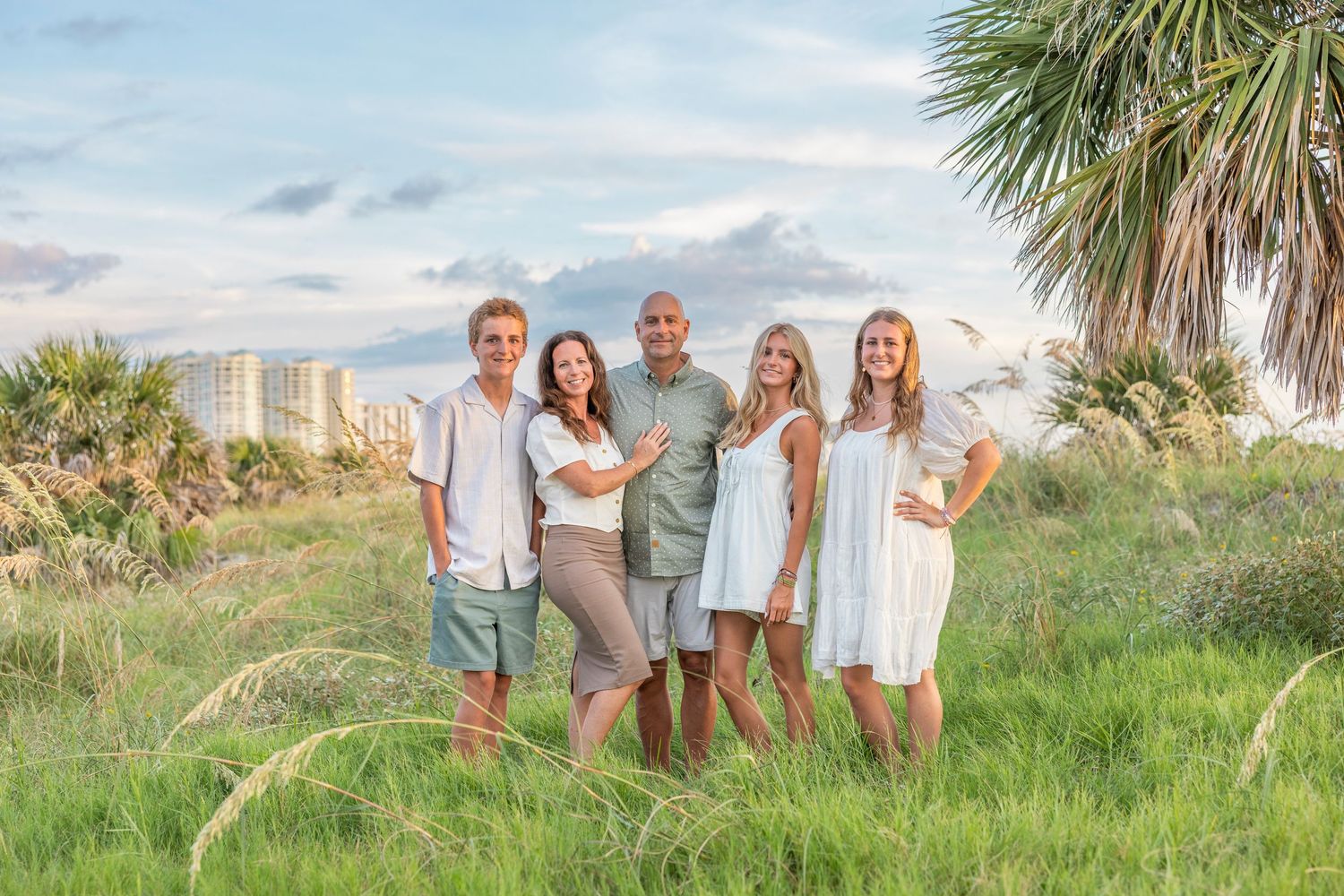 Family of five smiling together in a lush green coastal setting with palm trees and ocean buildings in the background.