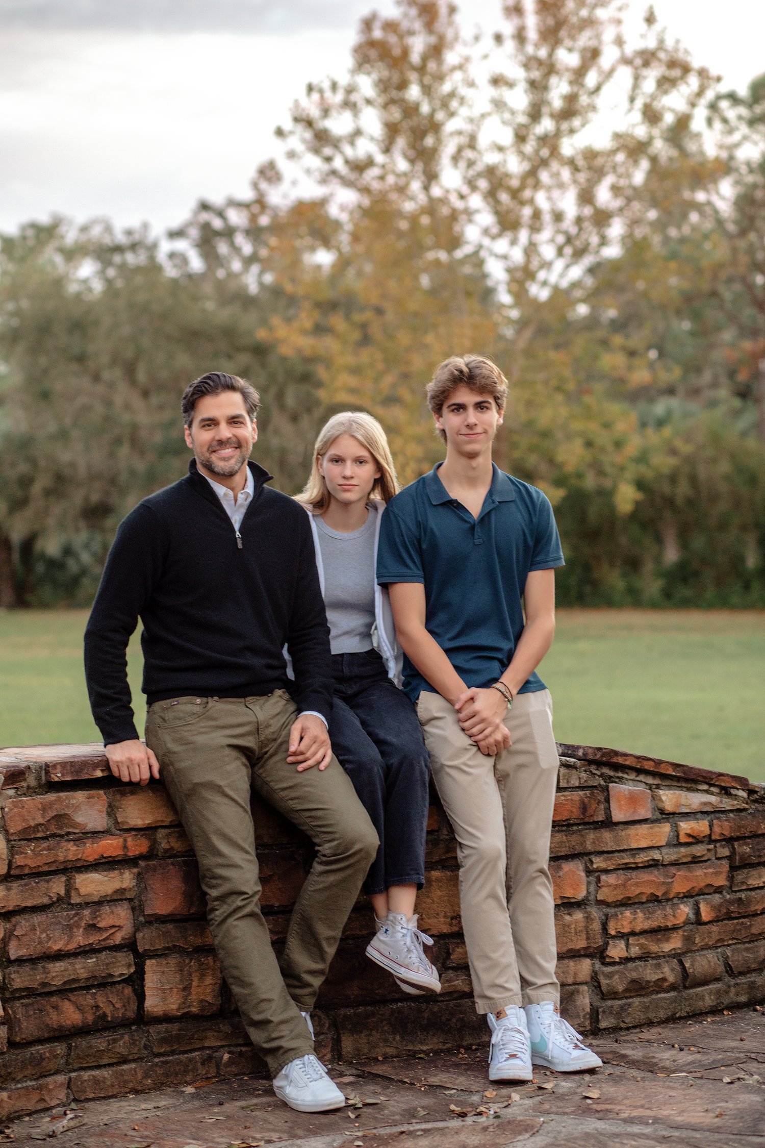 Family of three posing outdoors on a brick wall surrounded by lush greenery in casual attire.