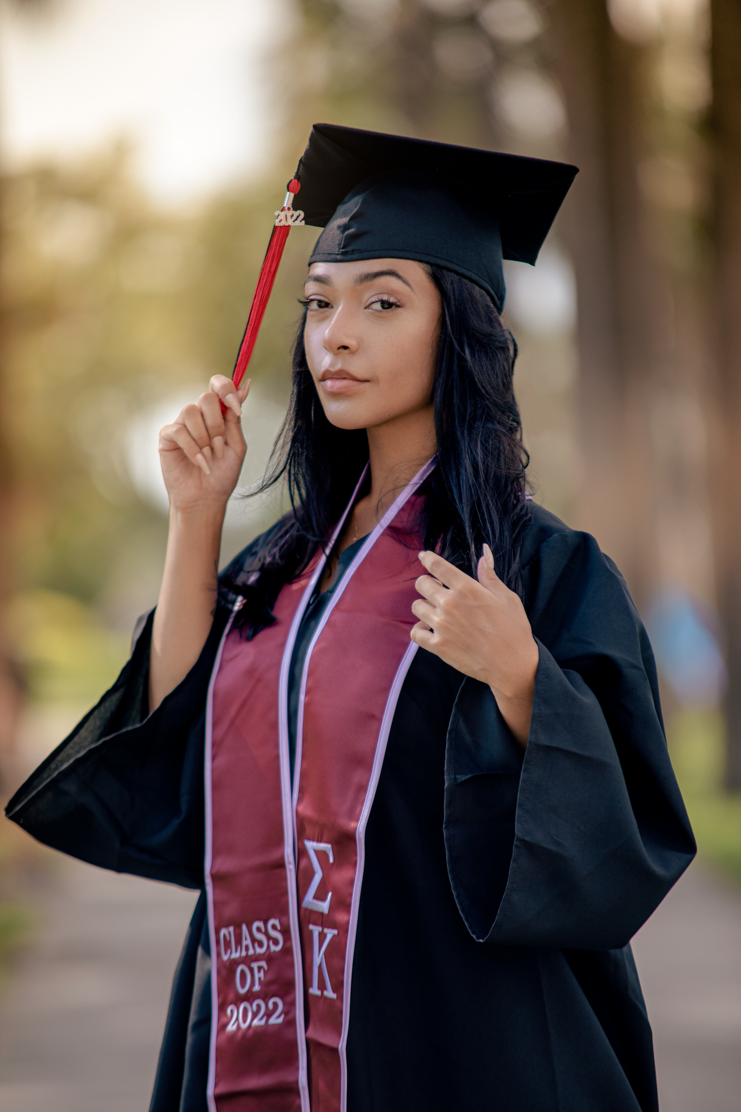 Young woman in black graduation gown and cap holding tassel, wearing Sigma Kappa Class of 2022 stole outdoors.