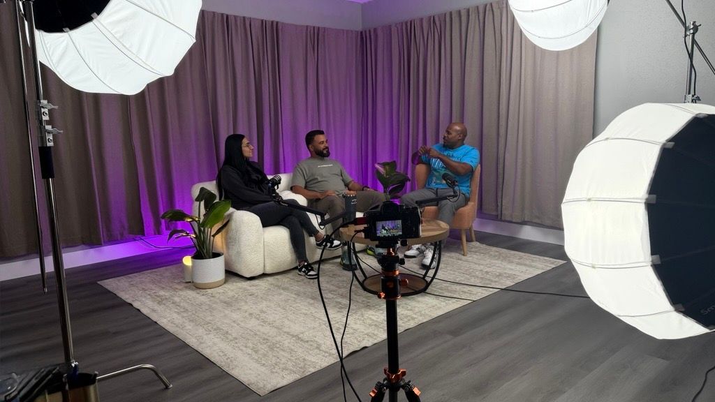 Three people seated in a professional TV studio setup with ring lights, cameras, and purple-lit curtain backdrop.