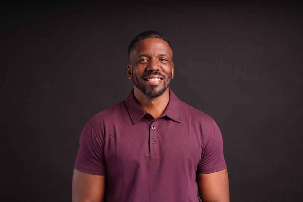 Smiling Black man in a maroon polo shirt posing against a dark background in a professional headshot photo.