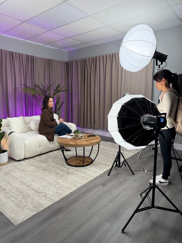 Photographer with umbrella light photographing a woman seated on a white sofa in a purple-lit studio.